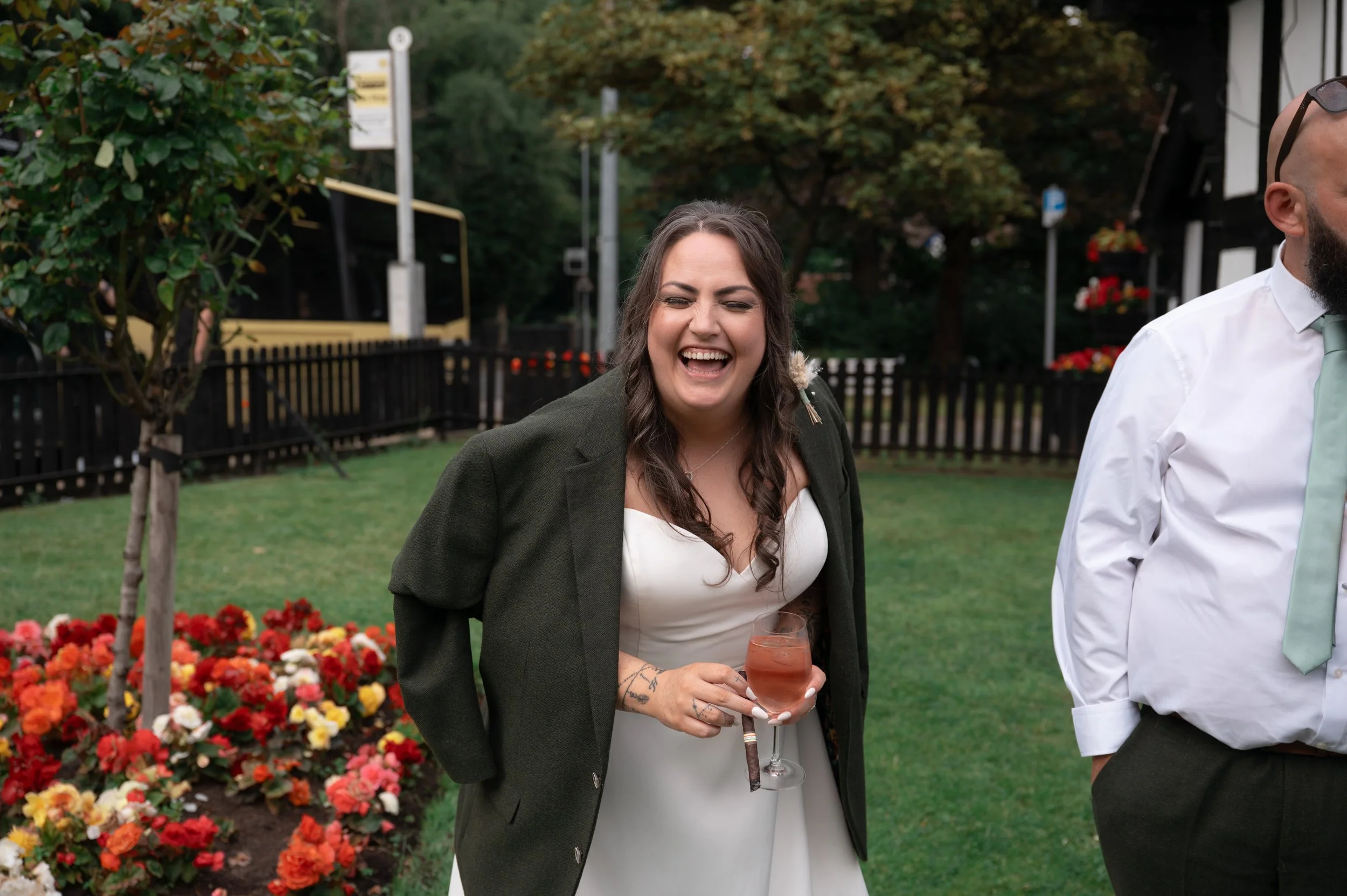 Smiling woman in white dress and green blazer holding a glass of pink-colored drink at an outdoor event, with a background of flowers, trees, and a black fence.