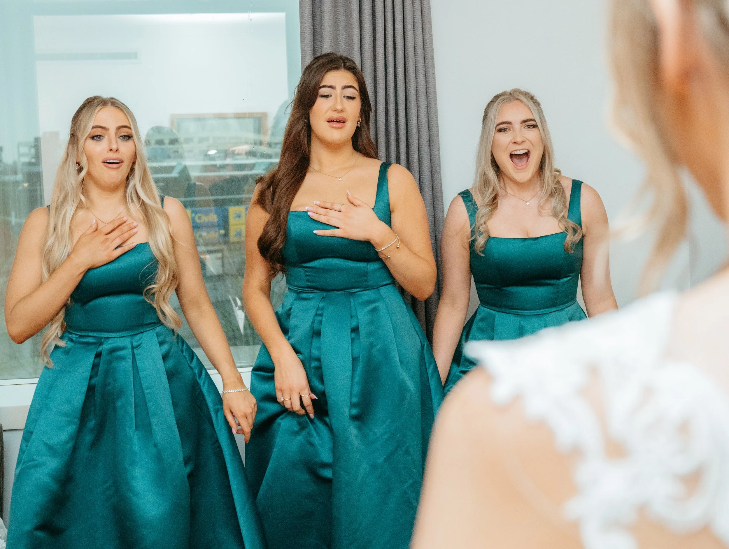 Three women in matching teal dresses reacting emotionally to a bride, who is partially visible on the right, at a wedding or special event.