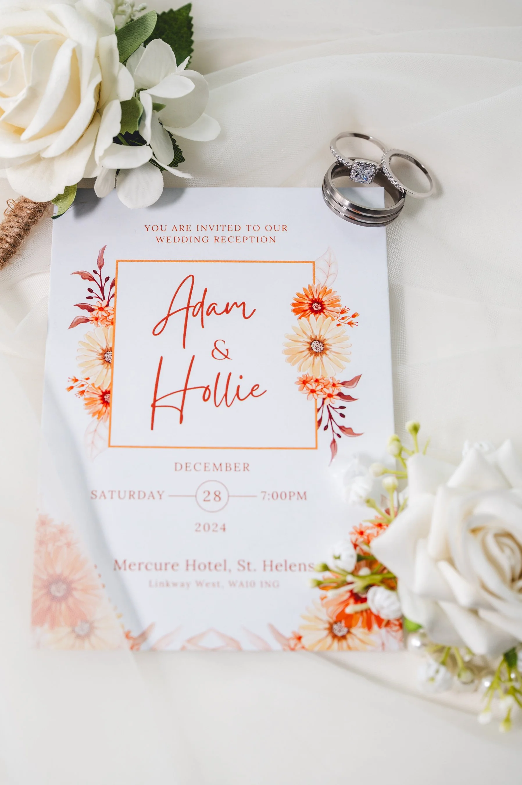 Wedding invitation card with floral border, rings, and flowers on a white fabric background.