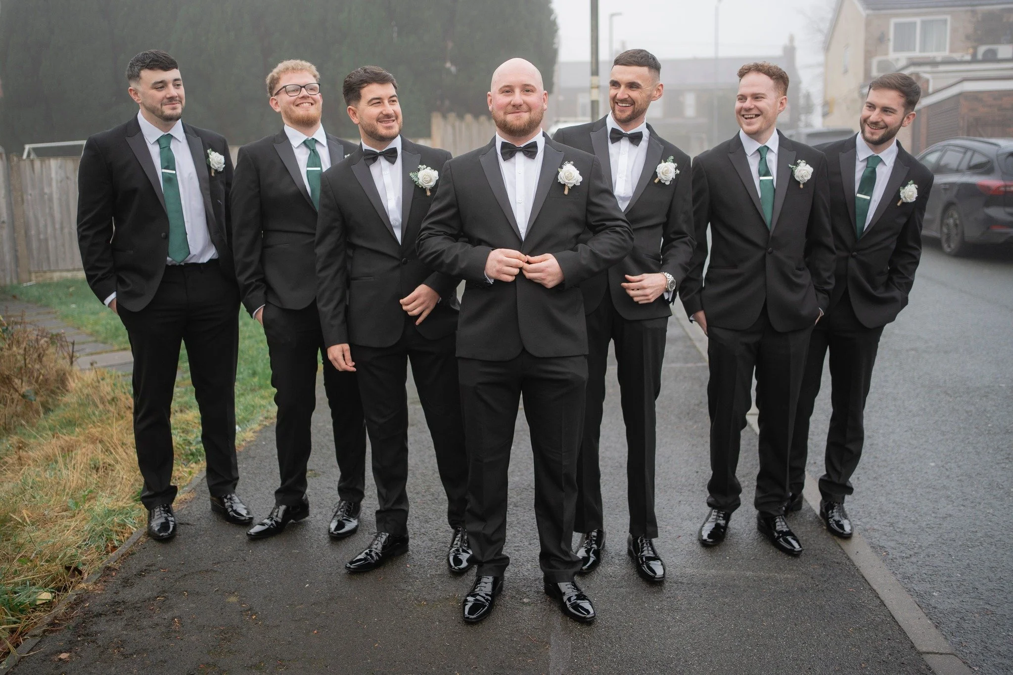A group of seven men dressed in black suits, white shirts, and green ties, standing outdoors on a cloudy day. The men appear to be part of a wedding party, with the man in front center wearing a tuxedo with a bowtie and boutonniere, smiling confident