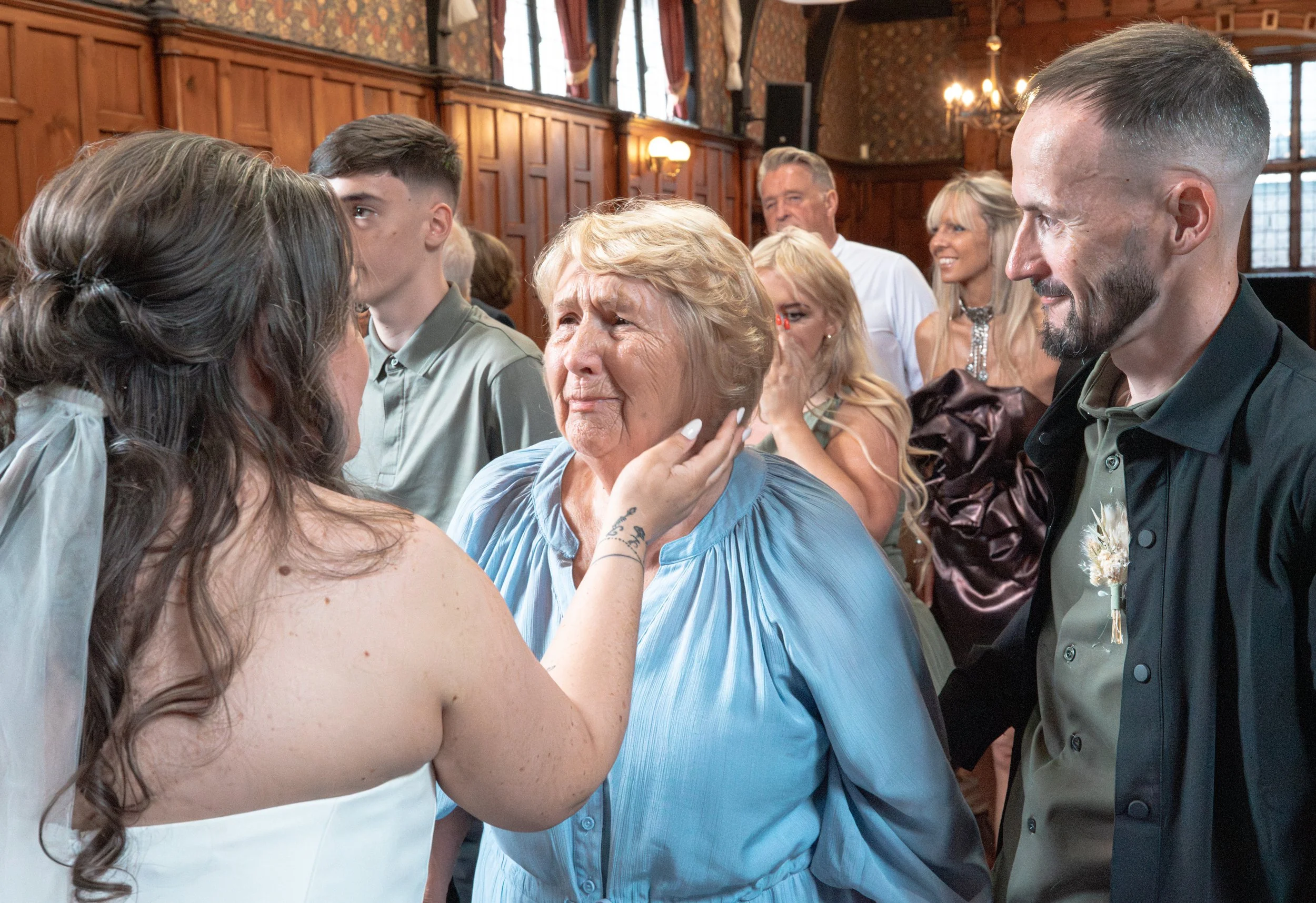 A woman in a wedding dress touches an older woman's face in an emotional scene during a wedding ceremony or celebration with multiple people present.