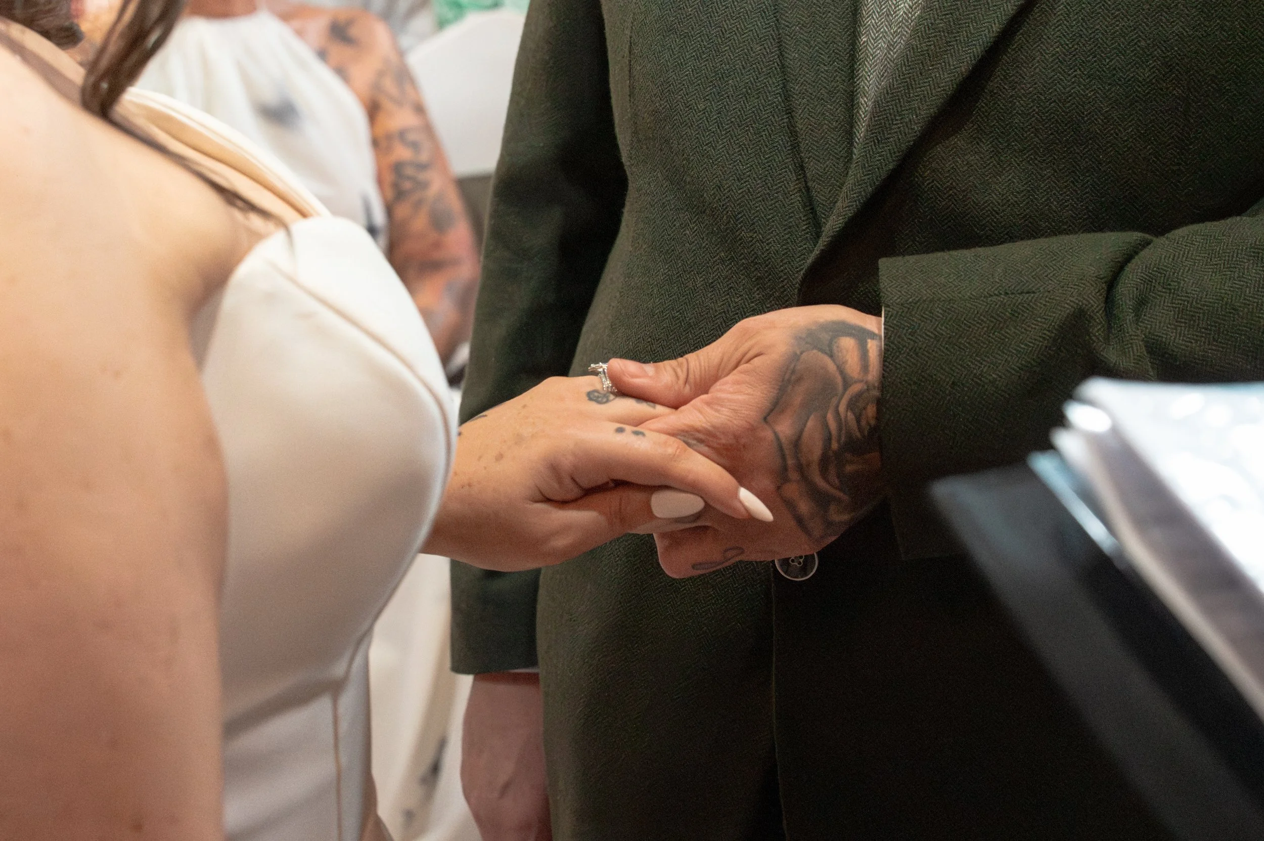 A close-up of a couple holding hands during a wedding ceremony, with the bride wearing a white dress and the groom in a dark green suit.