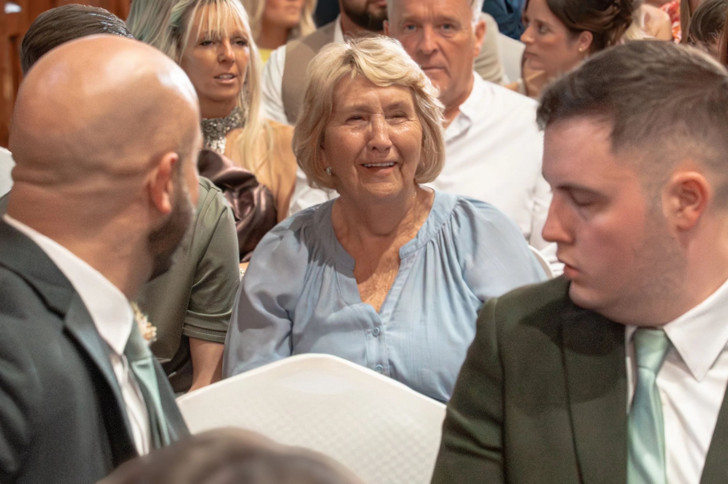 Elderly woman smiling and talking with two men in suits at a formal event, surrounded by other well-dressed people.