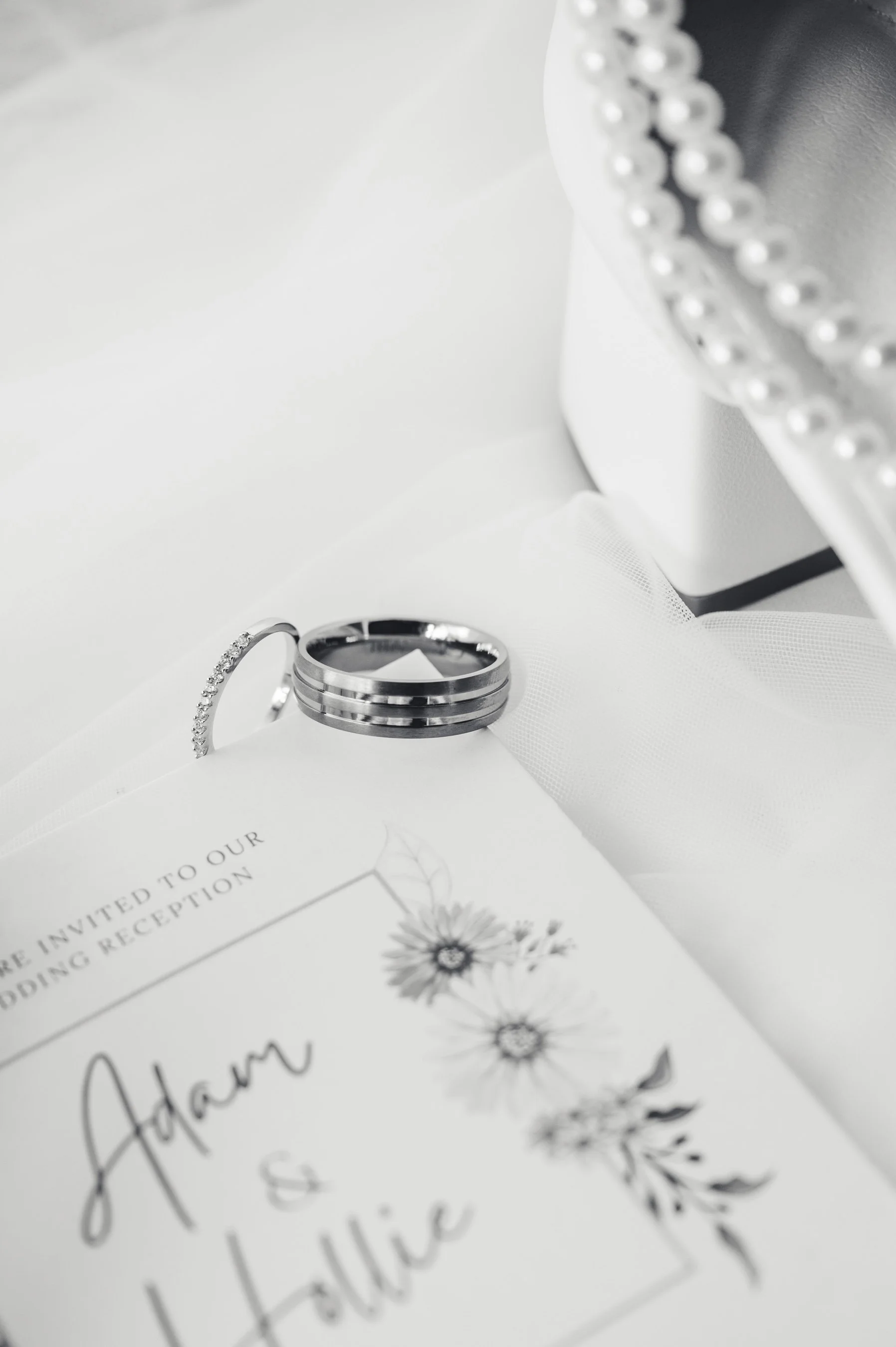 Wedding invitation with rings on top, featuring floral design and text reading "Adam & Julie," silver wedding bands, pearl-embellished jewelry, and white fabric background.