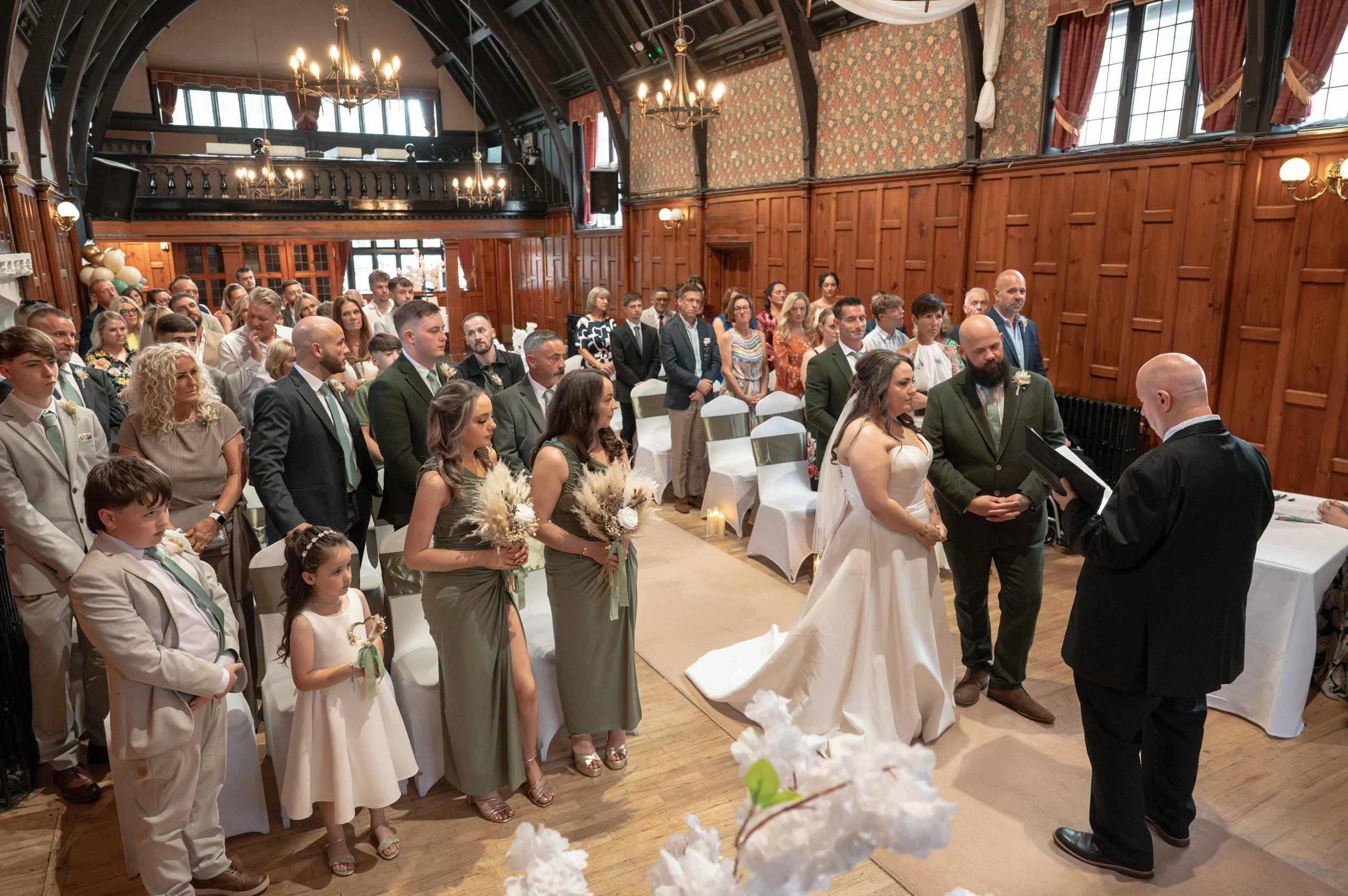 A wedding ceremony taking place in a historic, wood-paneled room with high ceilings, chandeliers, and tall windows. The bride and groom stand before an officiant, surrounded by their wedding party and guests seated or standing, dressed in formal atti