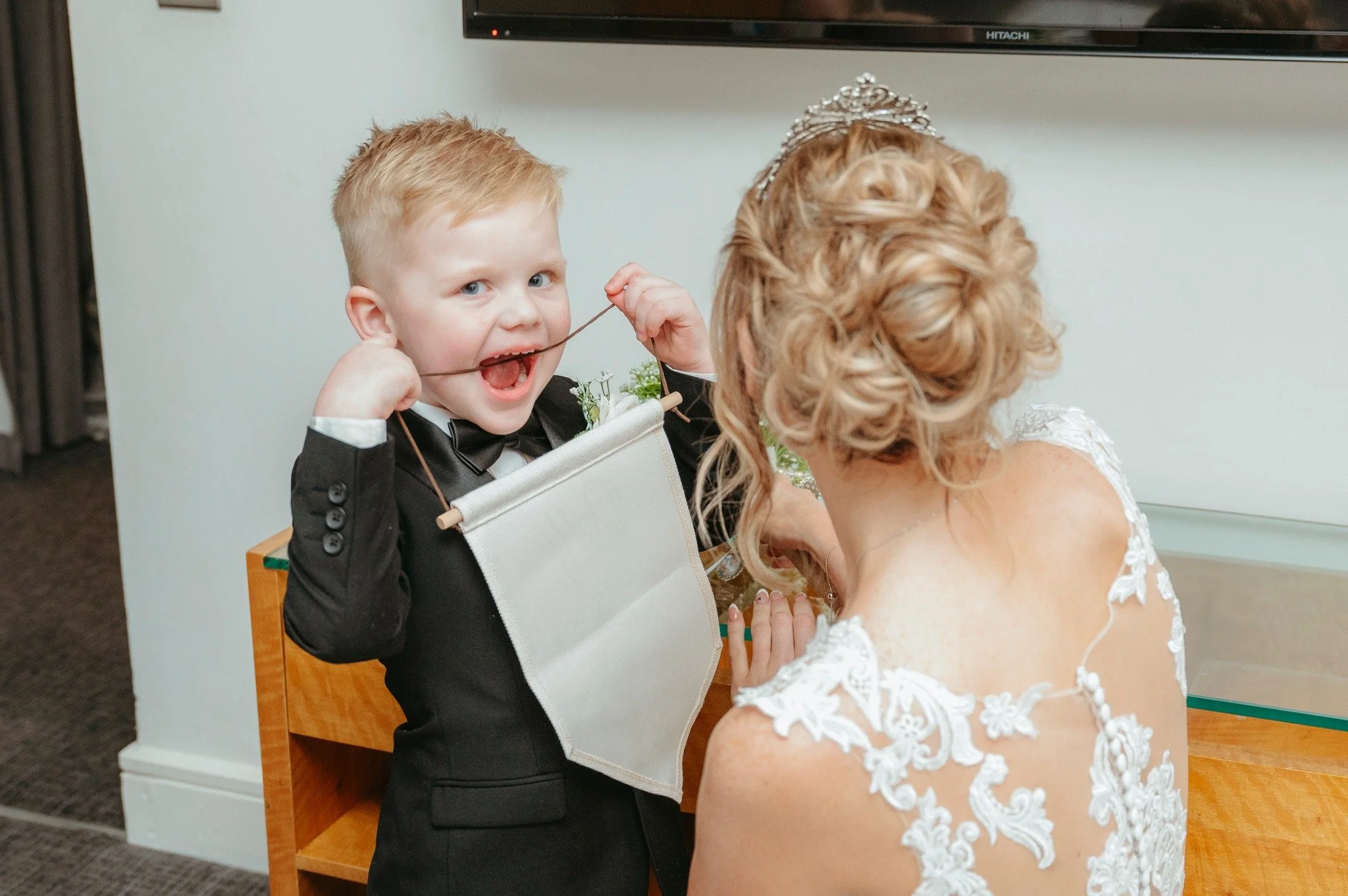 A young boy in a tuxedo playfully pulling on a string held by a woman in a wedding dress, smiling in a hotel room with a flat-screen TV in the background.