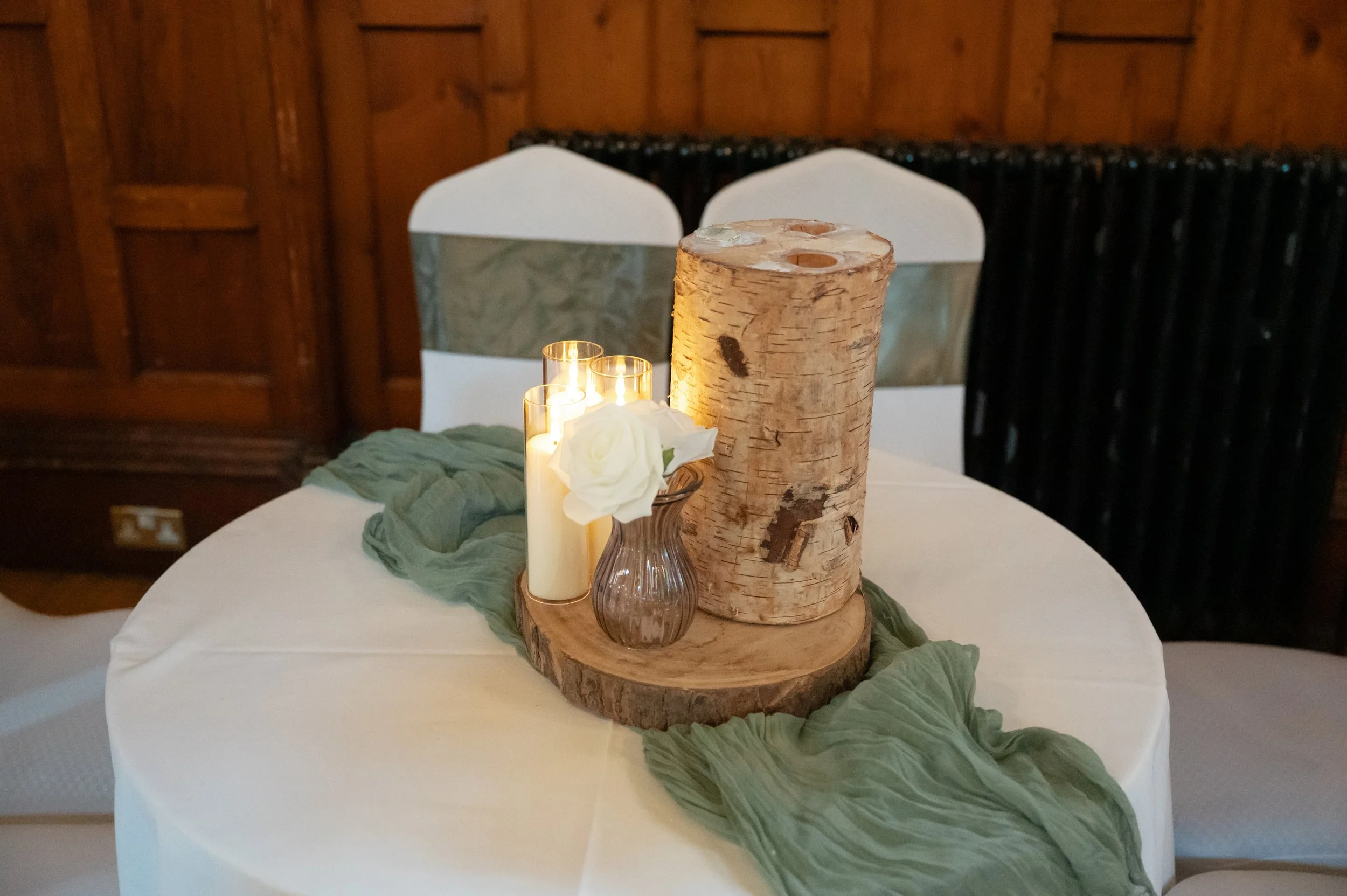 A round table decorated with a centerpiece consisting of a large birch bark candle holder, a small glass vase with white roses, a green fabric runner, and three lit candles. The table has white chairs with gold and white sashes in the background.