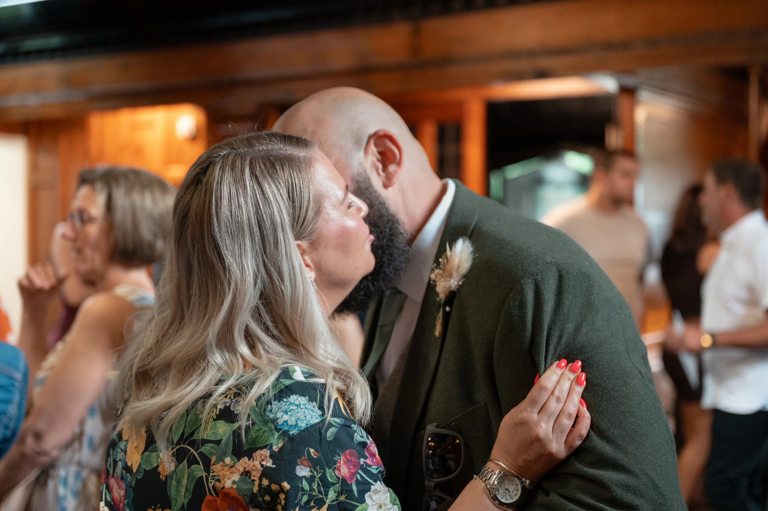 A couple is kissing at a social gathering. The woman has blonde hair, is wearing a floral dress, and a watch. The man is bald with a beard, wearing a dark blazer and a boutonnière. Other people are in the background, engaged in conversation.