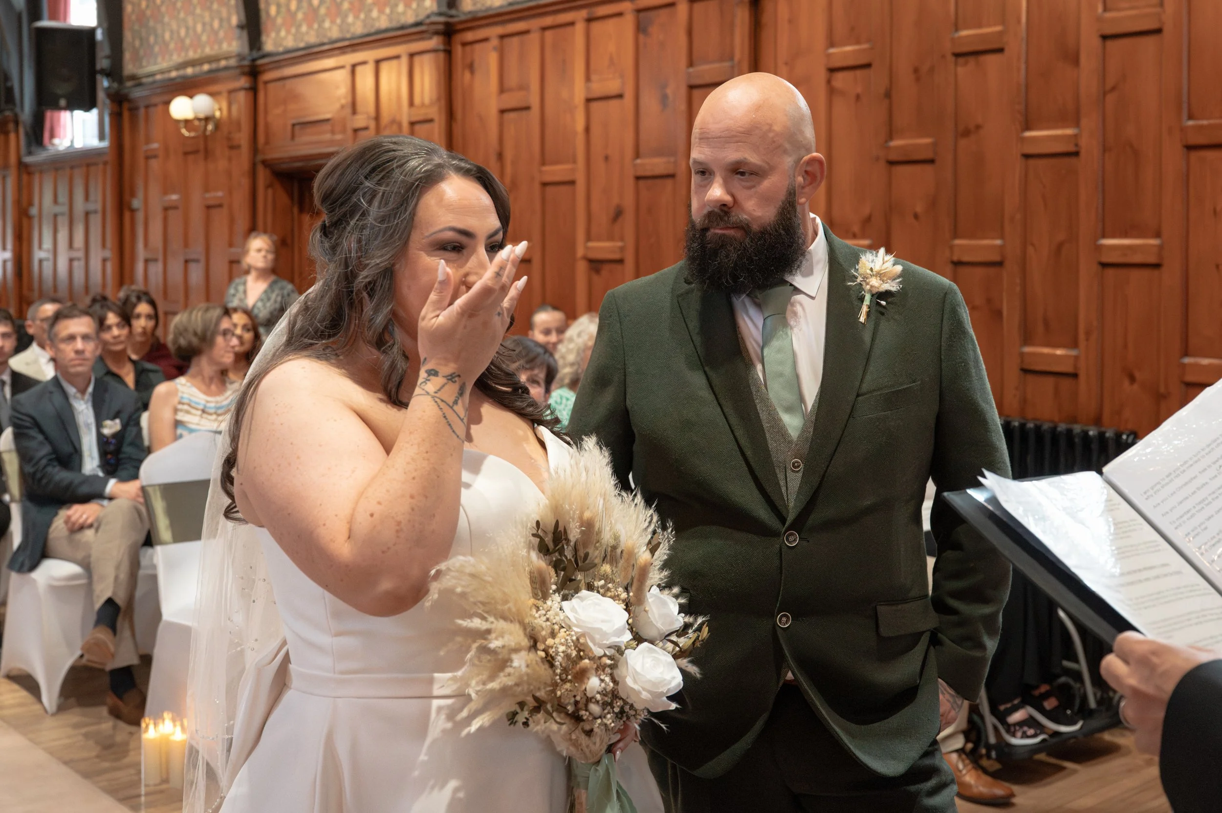 A bride and groom exchanging vows during a wedding ceremony in a wood-paneled room, with guests seated in the background.
