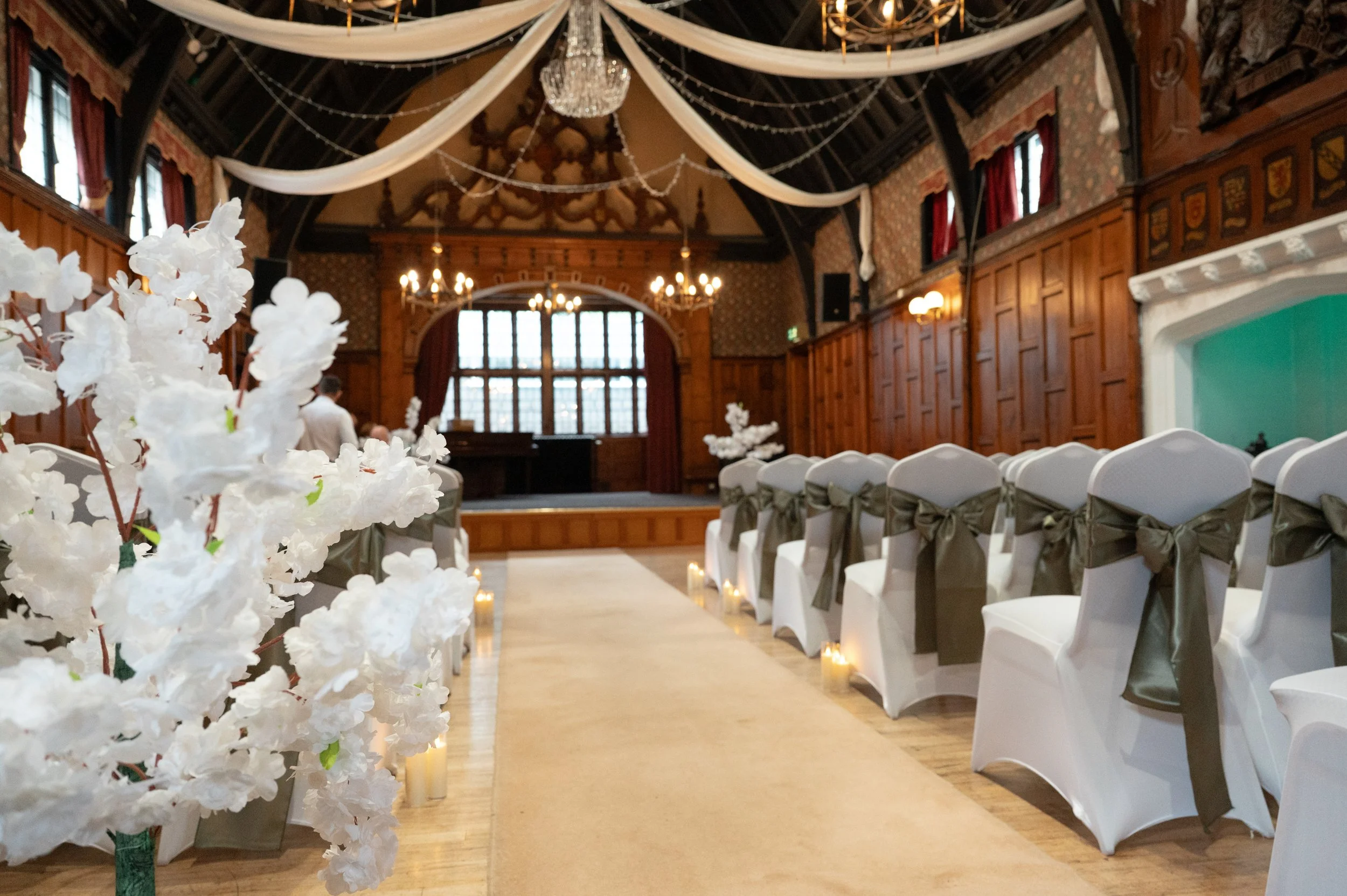Indoor wedding ceremony setup with white chairs decorated with dark green sashes, white flowers in the foreground, candles along the aisle, a stage with a live band, and elegant hanging drapes in a historic wooden hall.