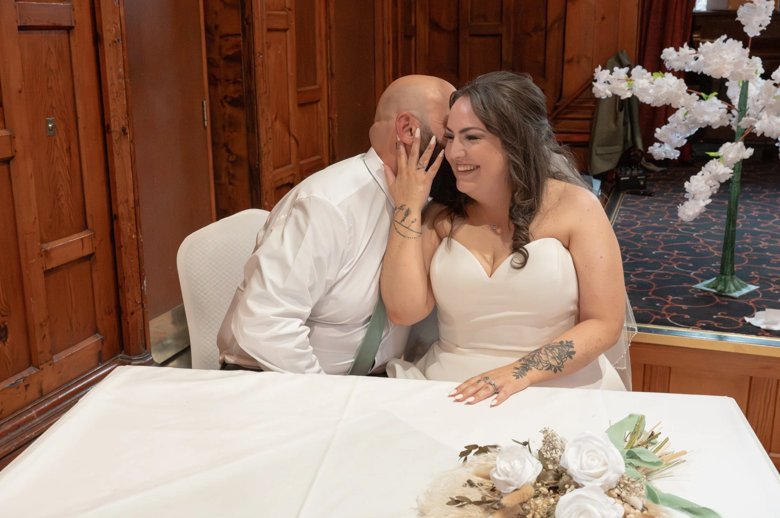 A newlywed couple sharing a joyful moment, with the bride touching the groom's face, woman smiling, in a warmly decorated room with wooden paneling, flowers, and wedding decor.