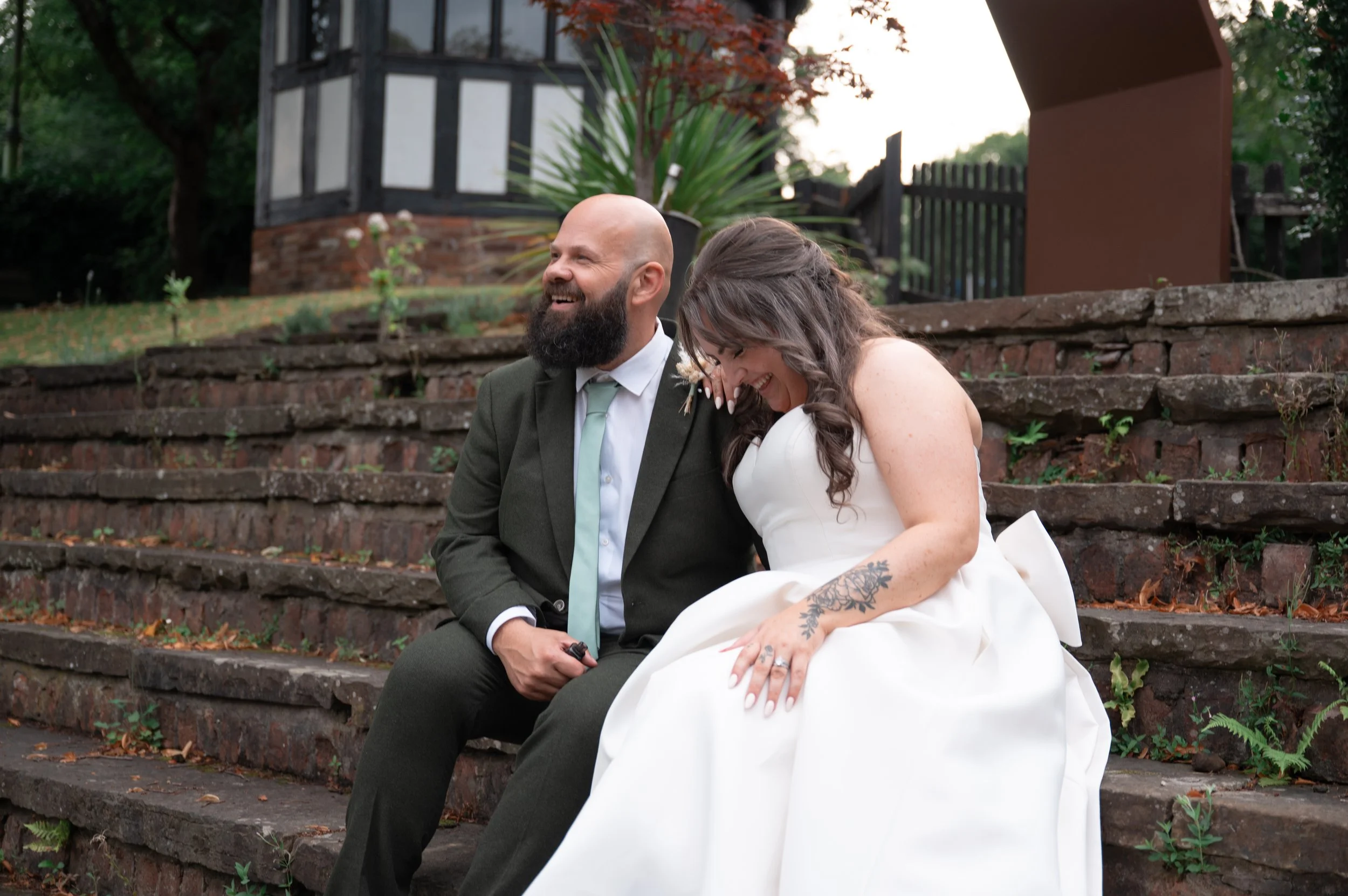 A newlywed couple sitting on outdoor brick steps, laughing and enjoying their wedding day. The groom has a beard and is wearing a dark suit with a light blue tie, while the bride is in a white wedding dress with a tattoo on her arm.
