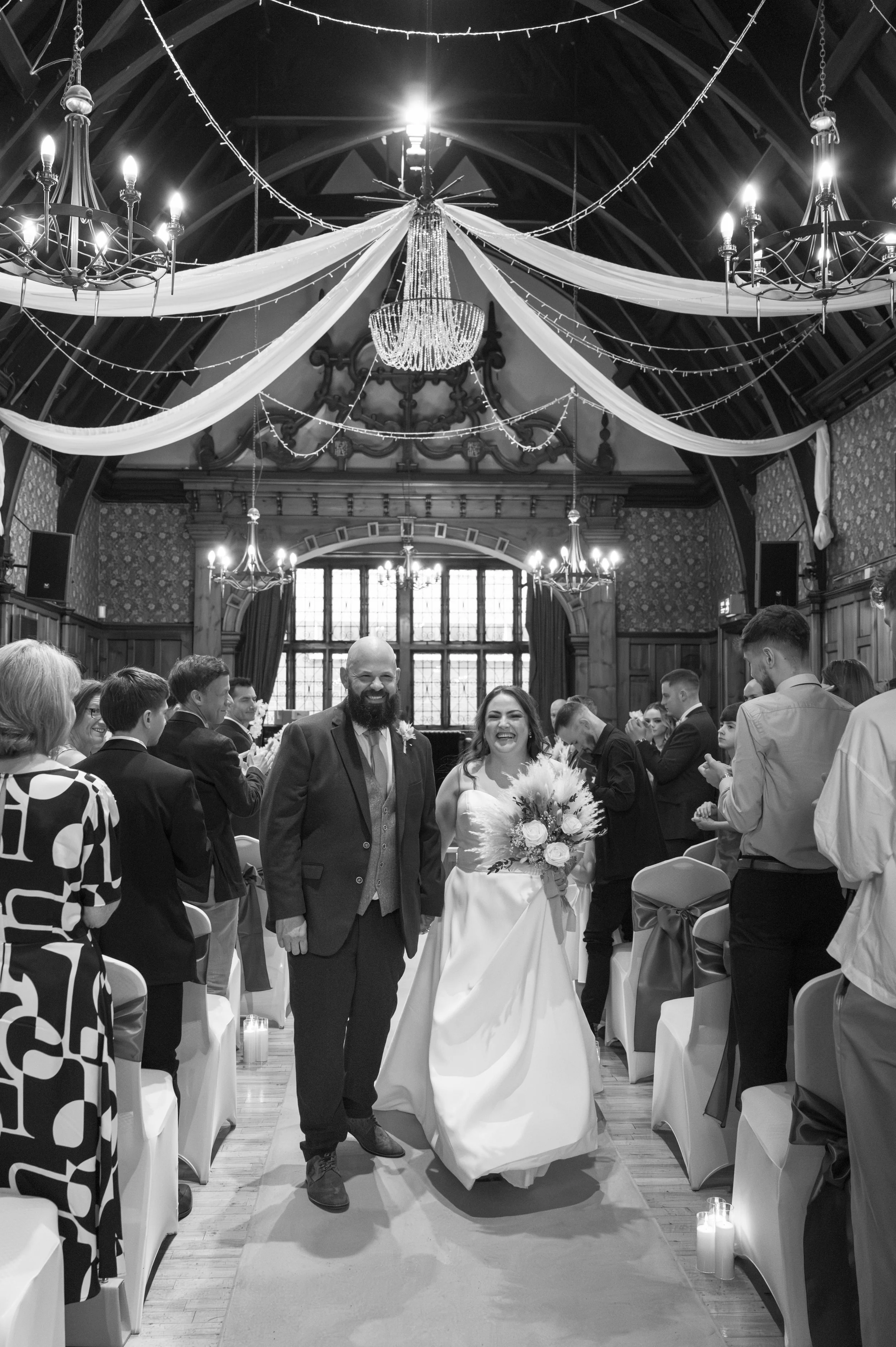 Black and white photo of a bride and groom walking down the aisle in a decorated wedding venue, surrounded by seated guests.