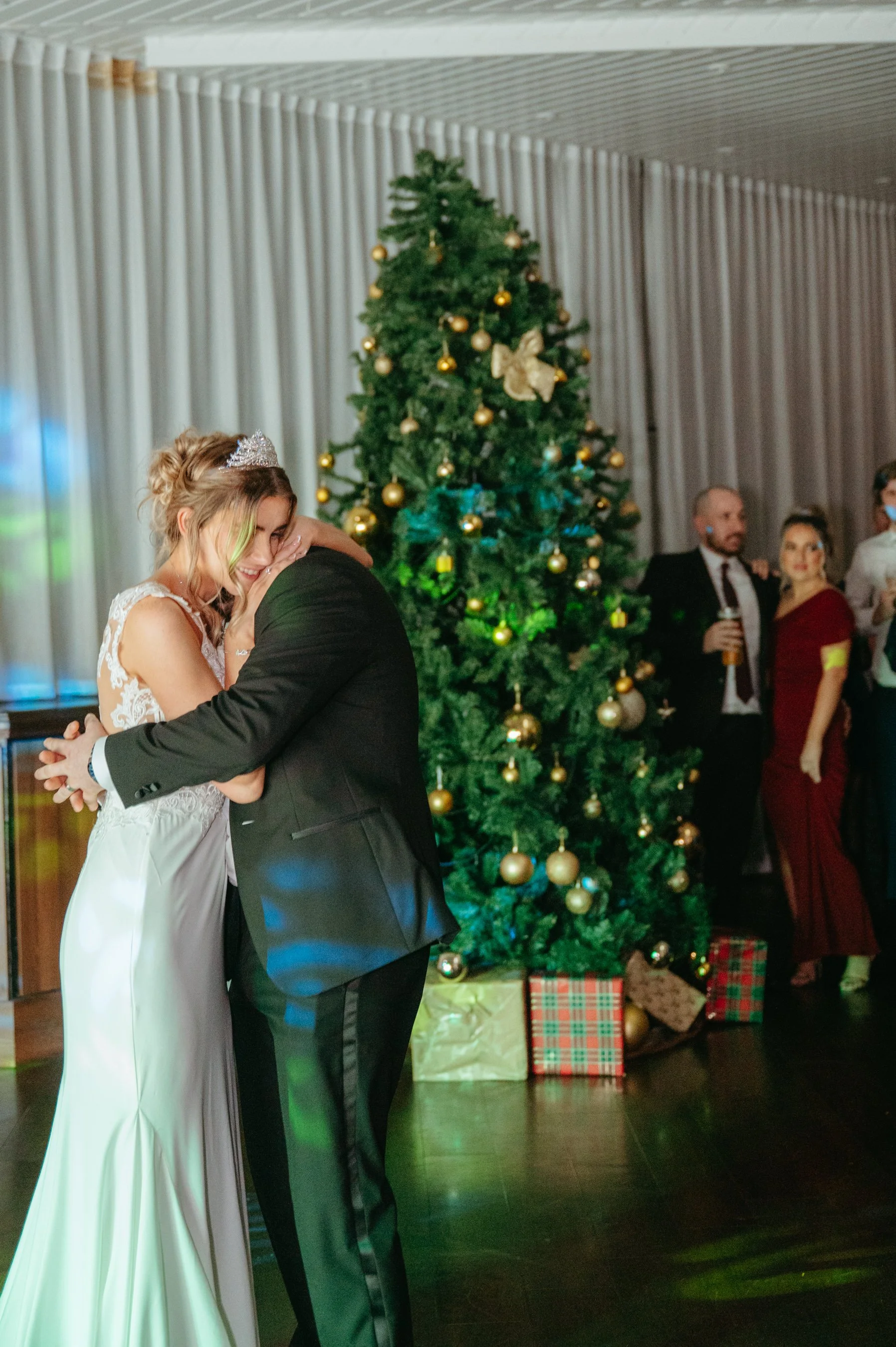 A couple embracing at a wedding reception next to a decorated Christmas tree with presents underneath.
