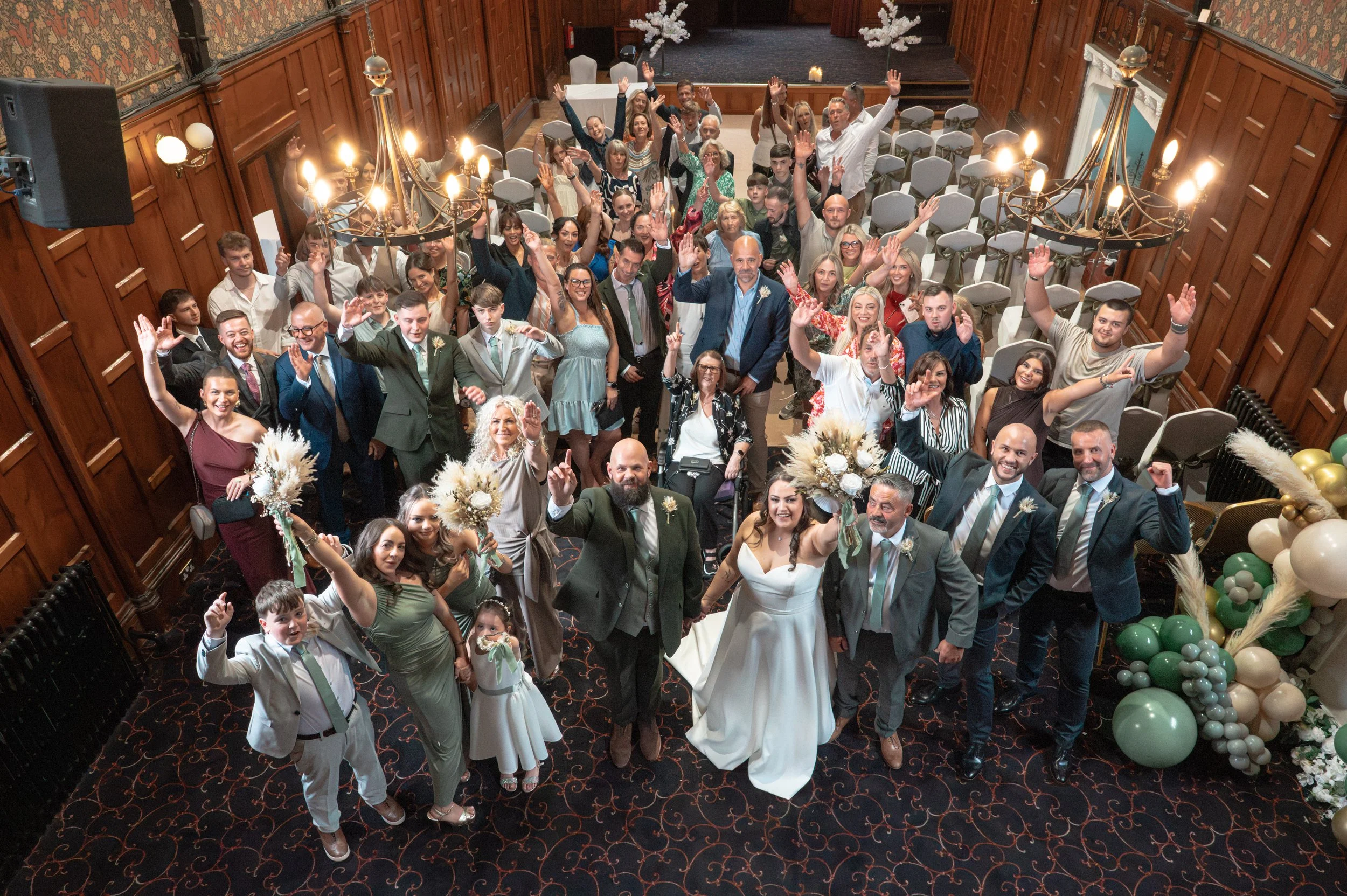 Crowd of wedding guests gathered in a decorated indoor venue, celebrating with the bride and groom at the front, holding flowers, and smiling at the camera.