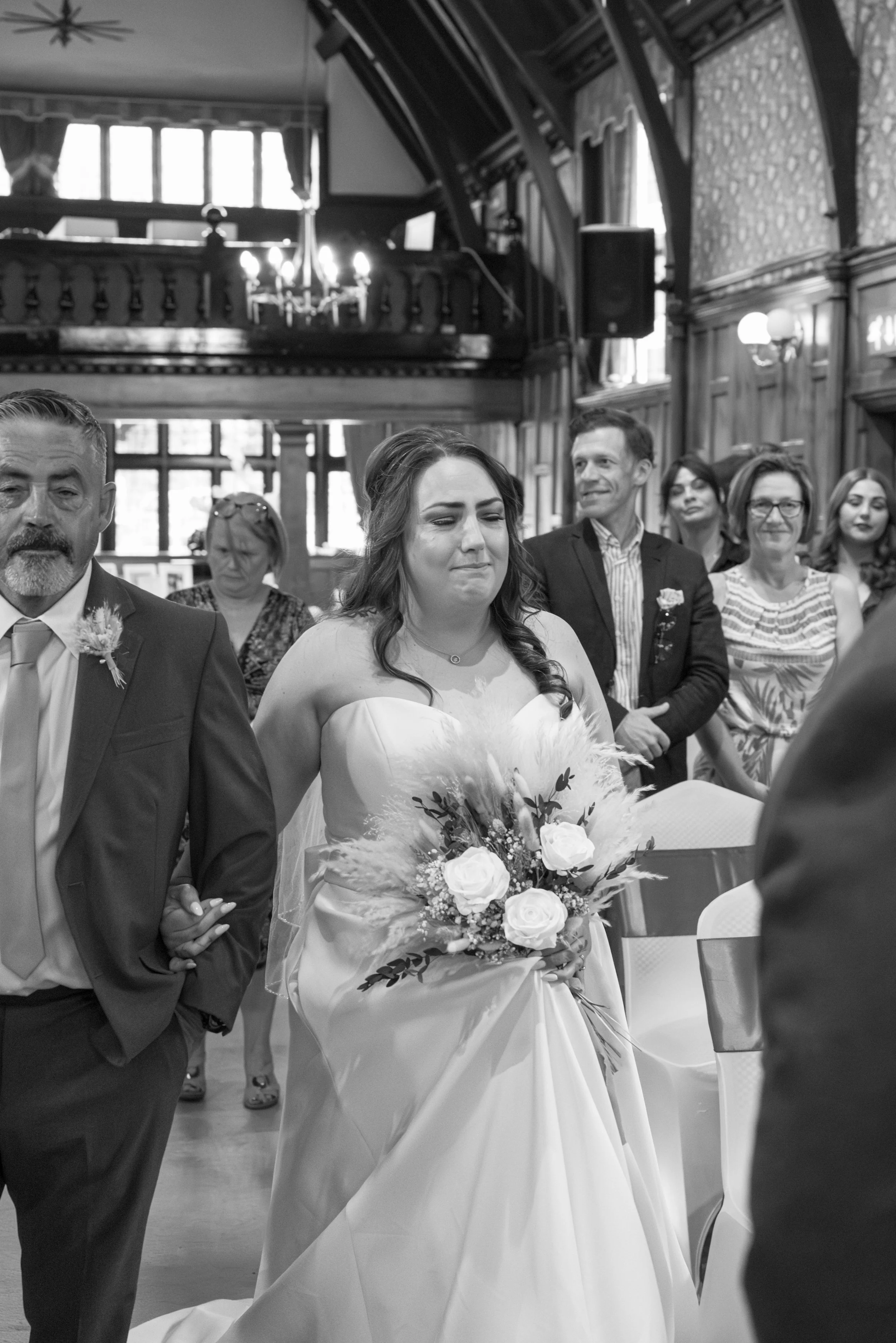 A bride with a distressed expression holding a bouquet of flowers, walking down the aisle with a man, surrounded by wedding guests in a decorated wooden interior.