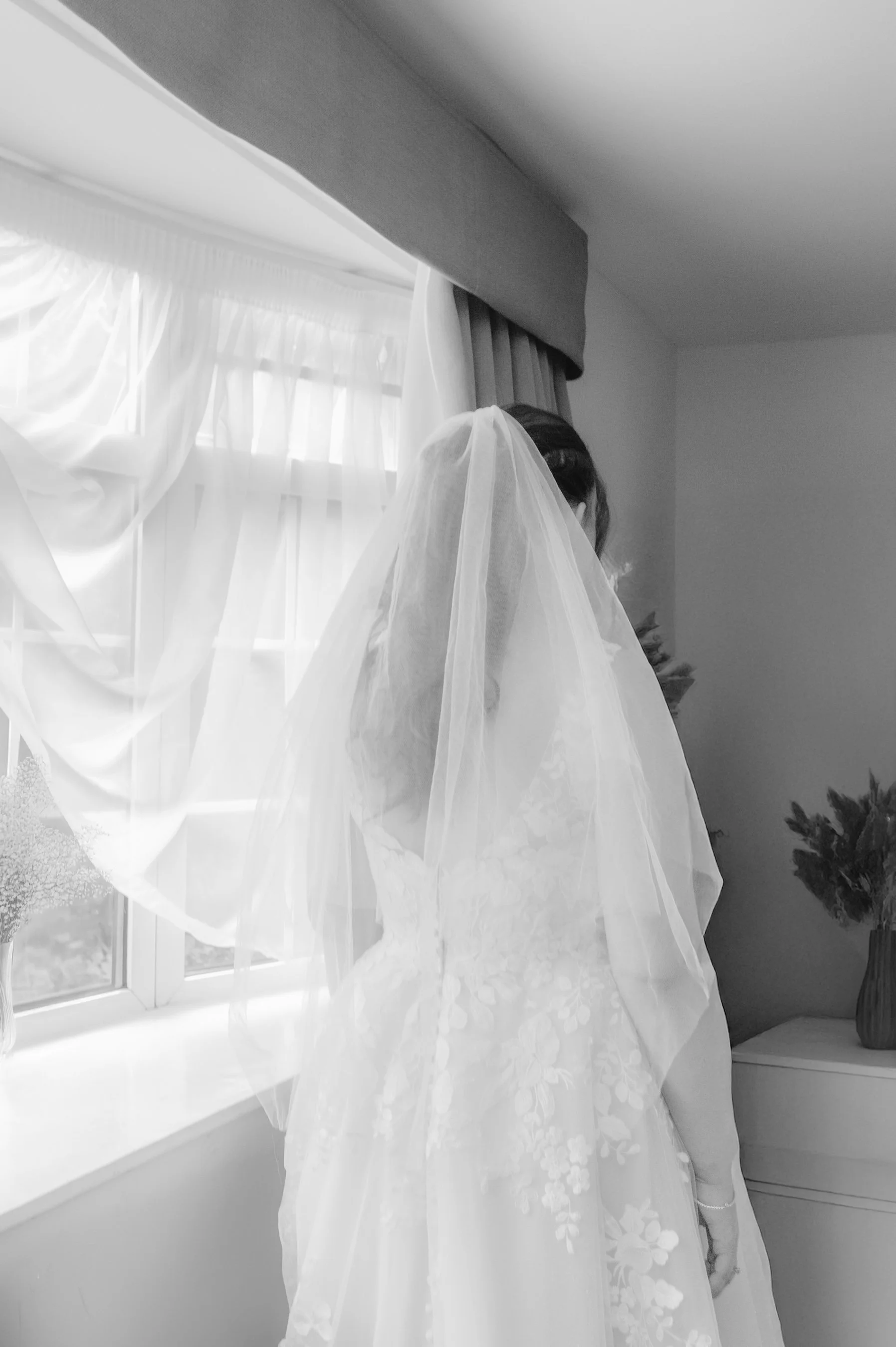A bride in a wedding gown and veil standing near a window with curtains, facing away from the camera.