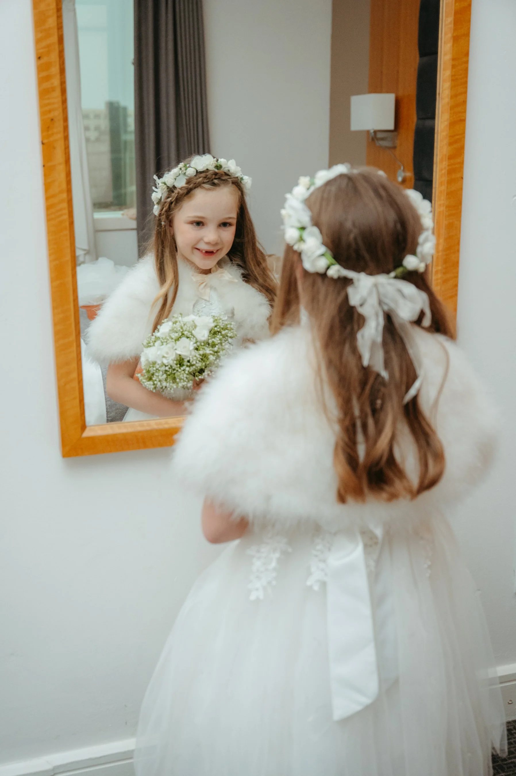 A young girl in a white dress with a floral headband and faux fur shawl, holding a bouquet, looking at her reflection in a mirror.