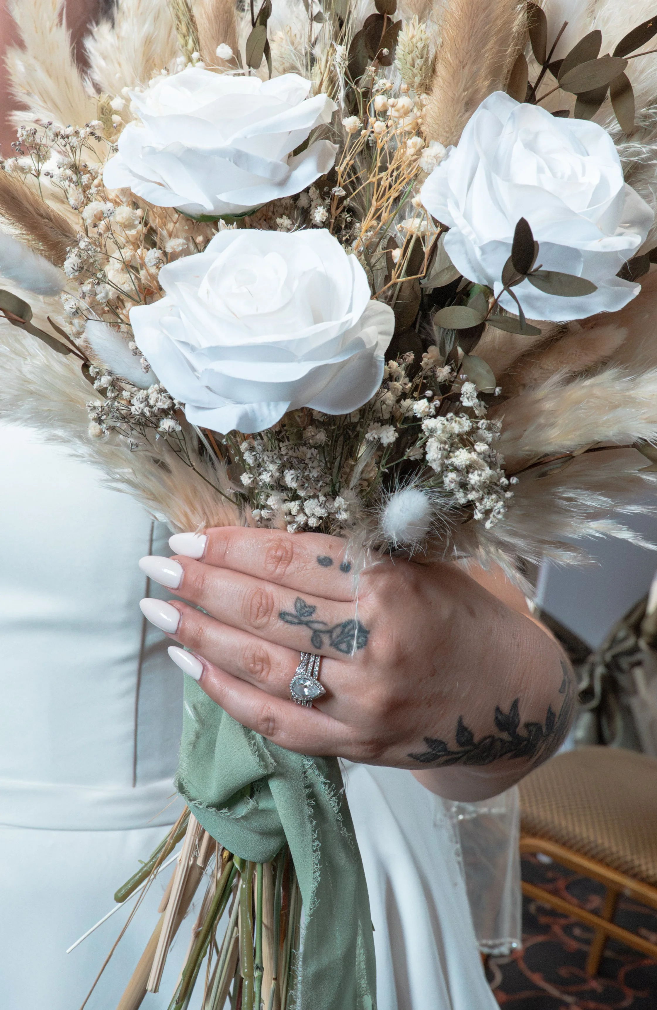 A person holding a bouquet of white roses, dried flowers, and beige pampas grass, with visible tattoos on their fingers and hand, wearing rings and polished white nails.