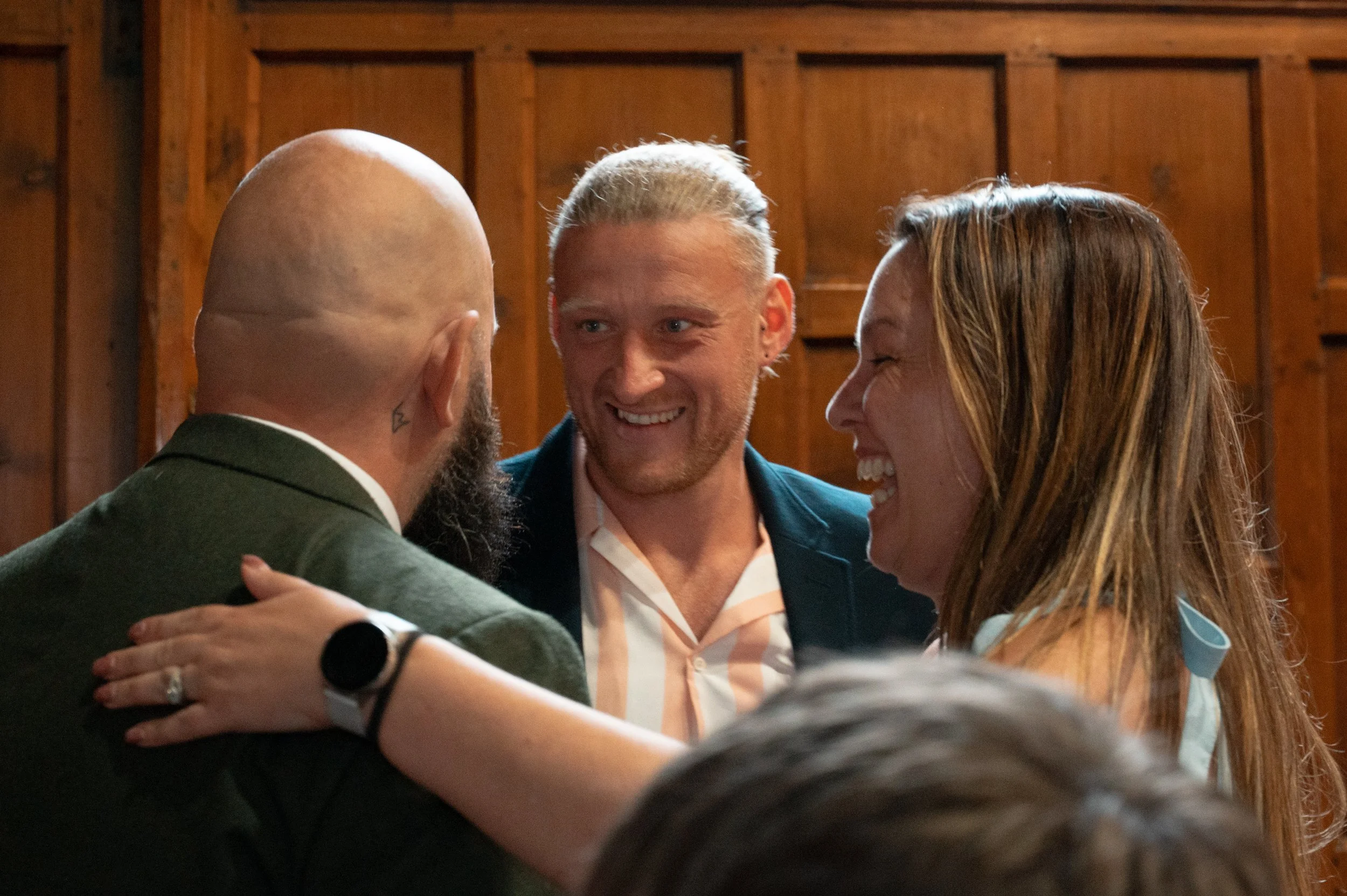 Three people smiling and hugging at an indoor gathering with wooden walls in the background.