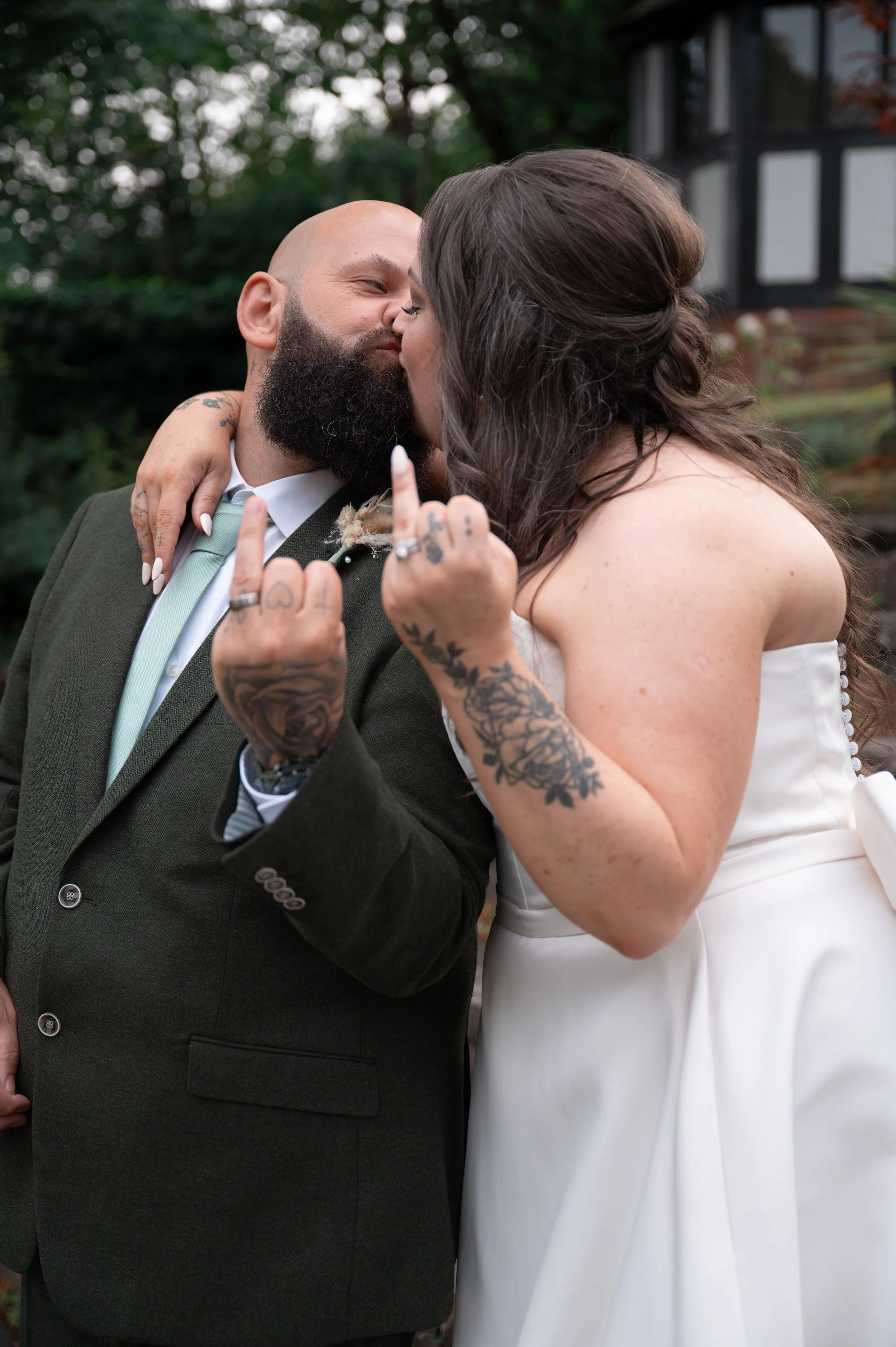 A couple kissing outdoors, with the woman in a white wedding dress and the man in a dark suit, both showing tattoos and making a middle finger gesture.