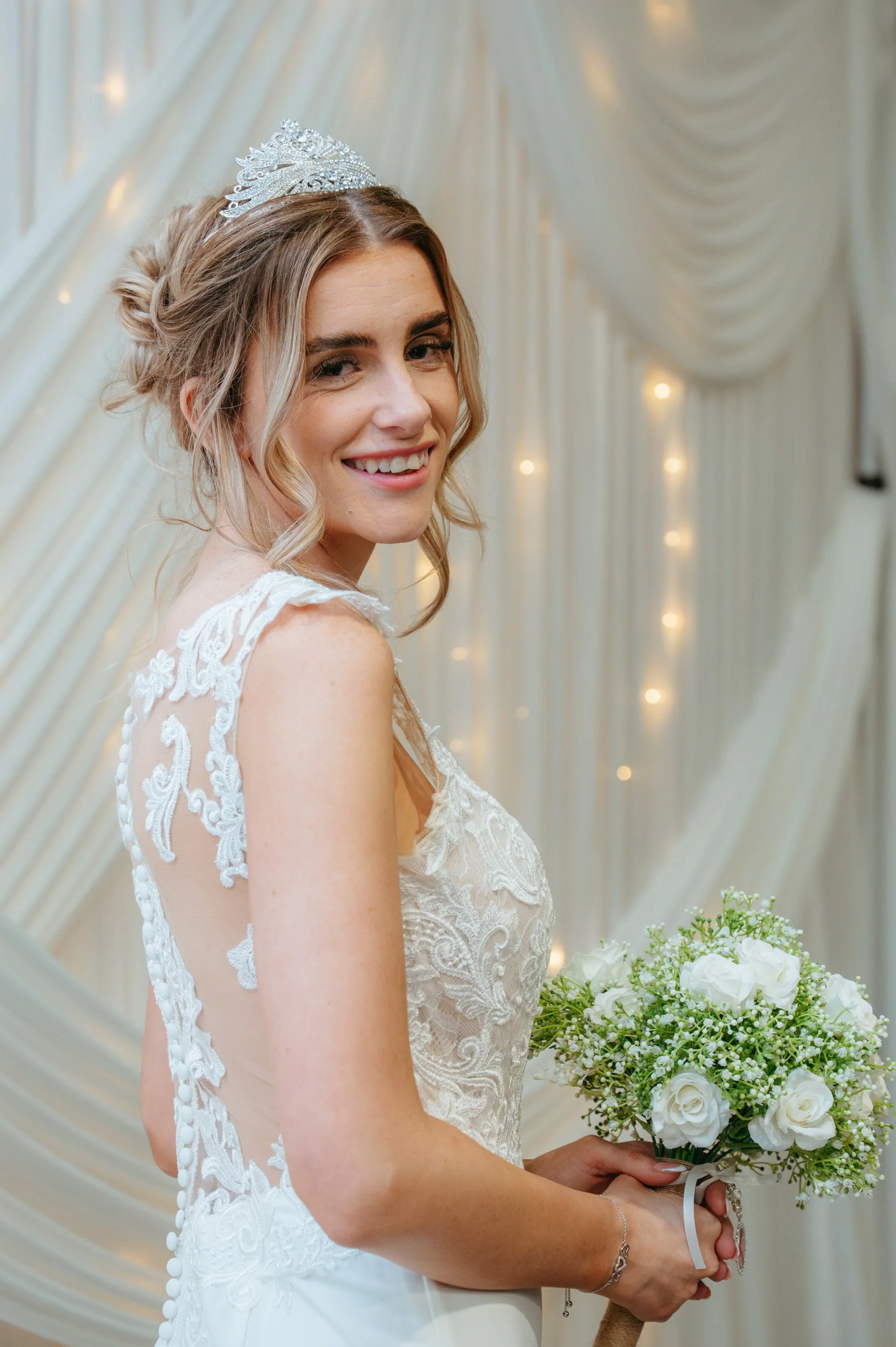 Bride in a white lace wedding dress holds a bouquet of white roses and baby's breath, smiling with a tiara in her styled hair, standing against a draped, fairy-lit background.