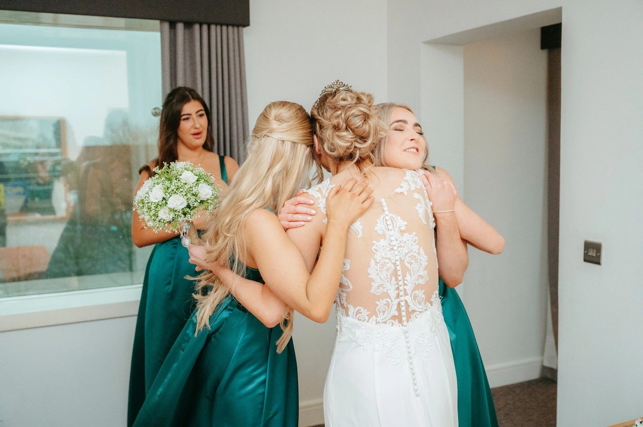 A bride is hugging three women, one with long blonde hair, one with short curly blonde hair, and one in a teal dress, in a room with white walls. One of the women in teal is holding a bouquet of white roses and greenery, and another woman in teal is
