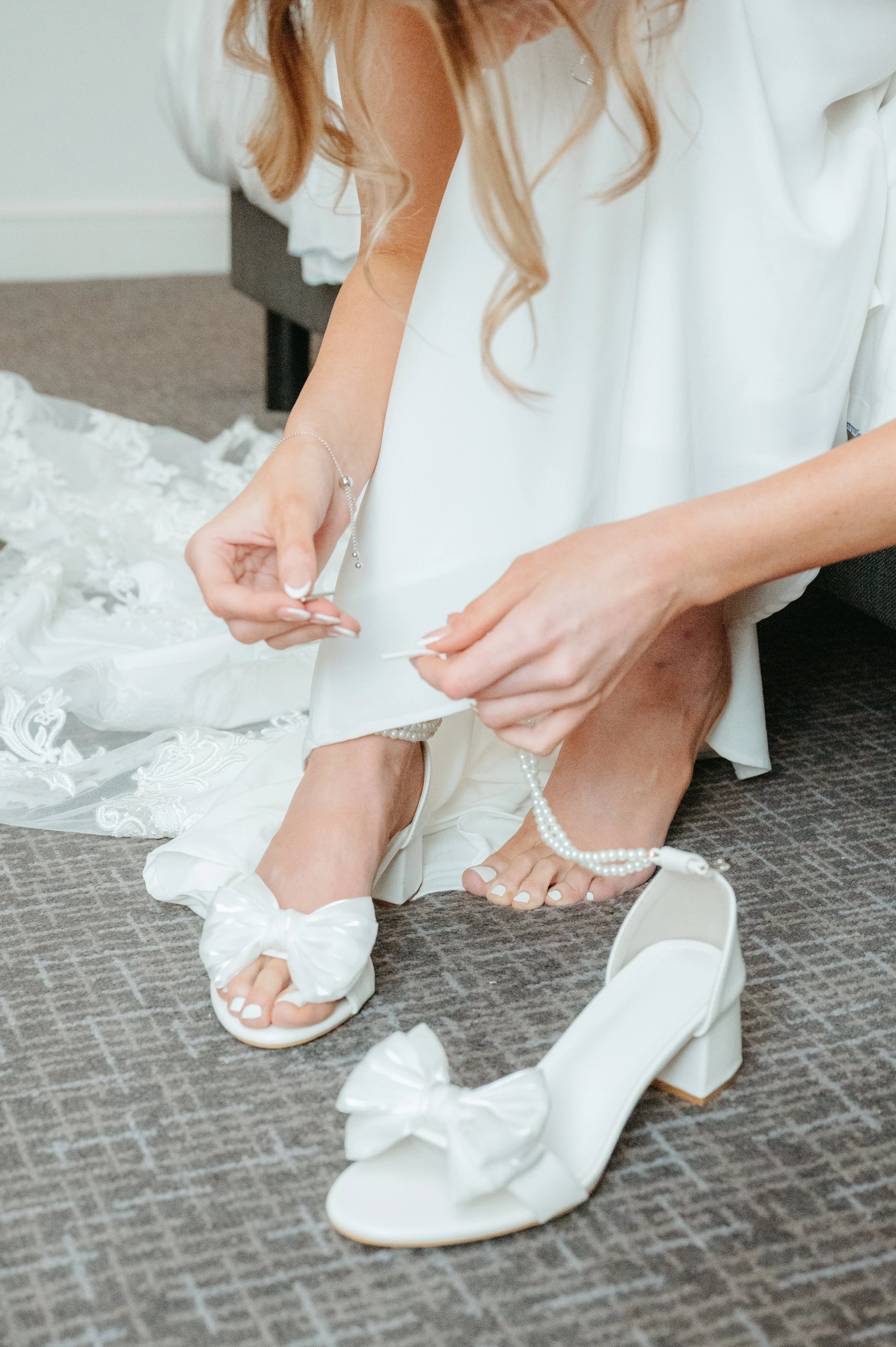 Bride wearing white slippers with bows, adjusting her wedding dress on a grey carpeted floor