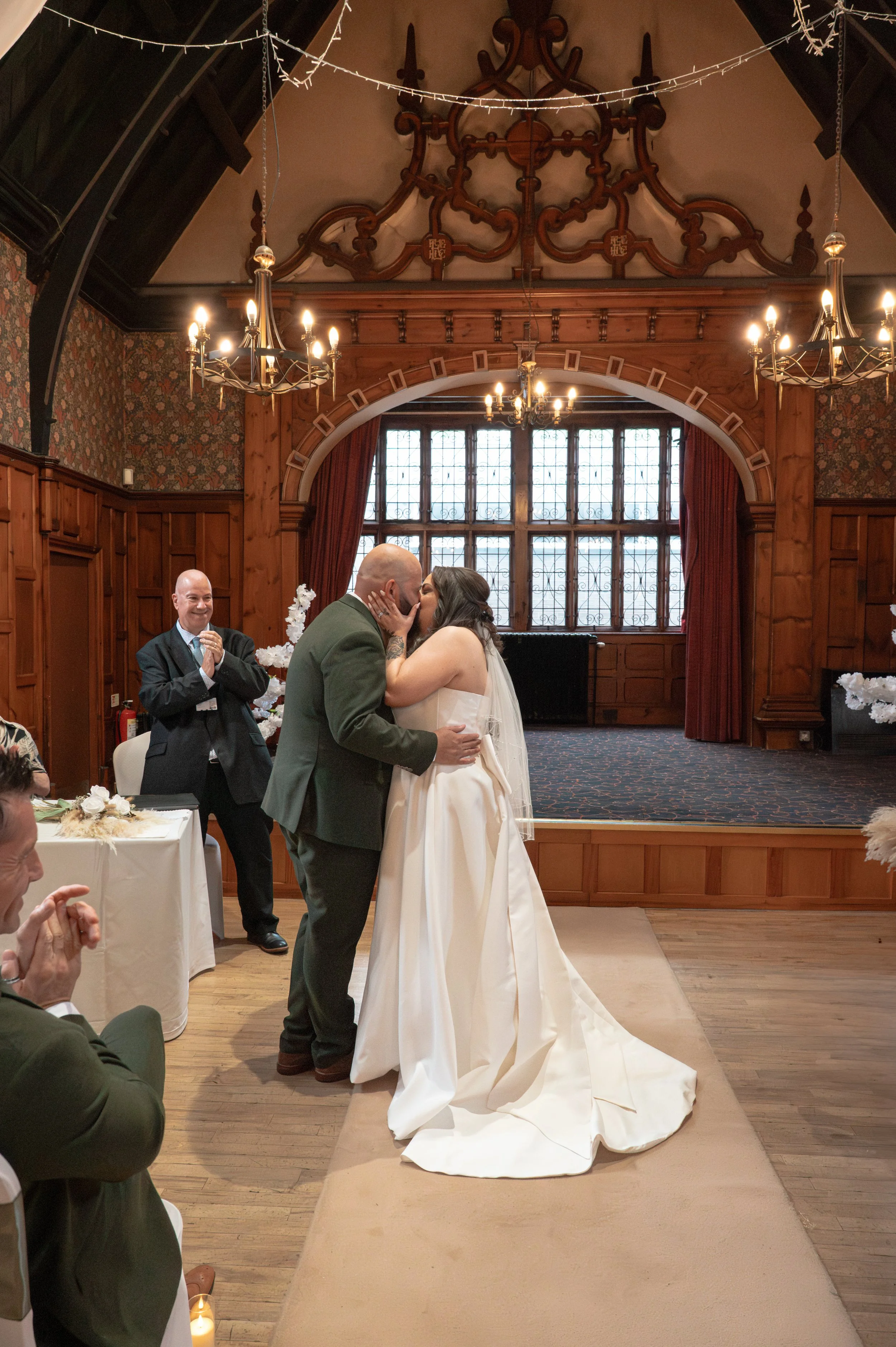 A wedding ceremony with a bride and groom sharing a kiss at the altar inside a wood-paneled room with large window, chandeliers, and guests watching.