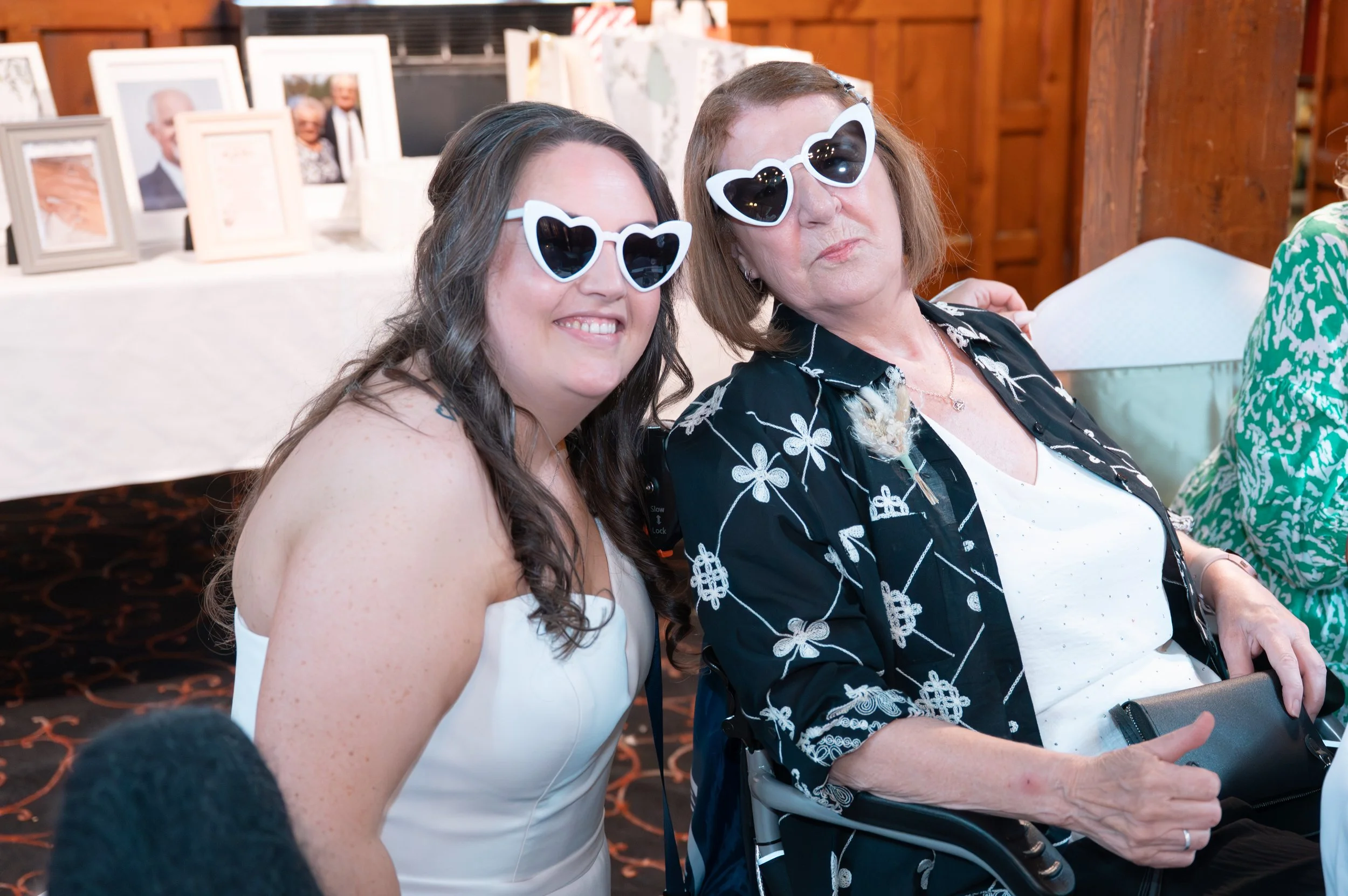Two women wearing heart-shaped sunglasses sitting together at a social event, smiling and posing for the camera.