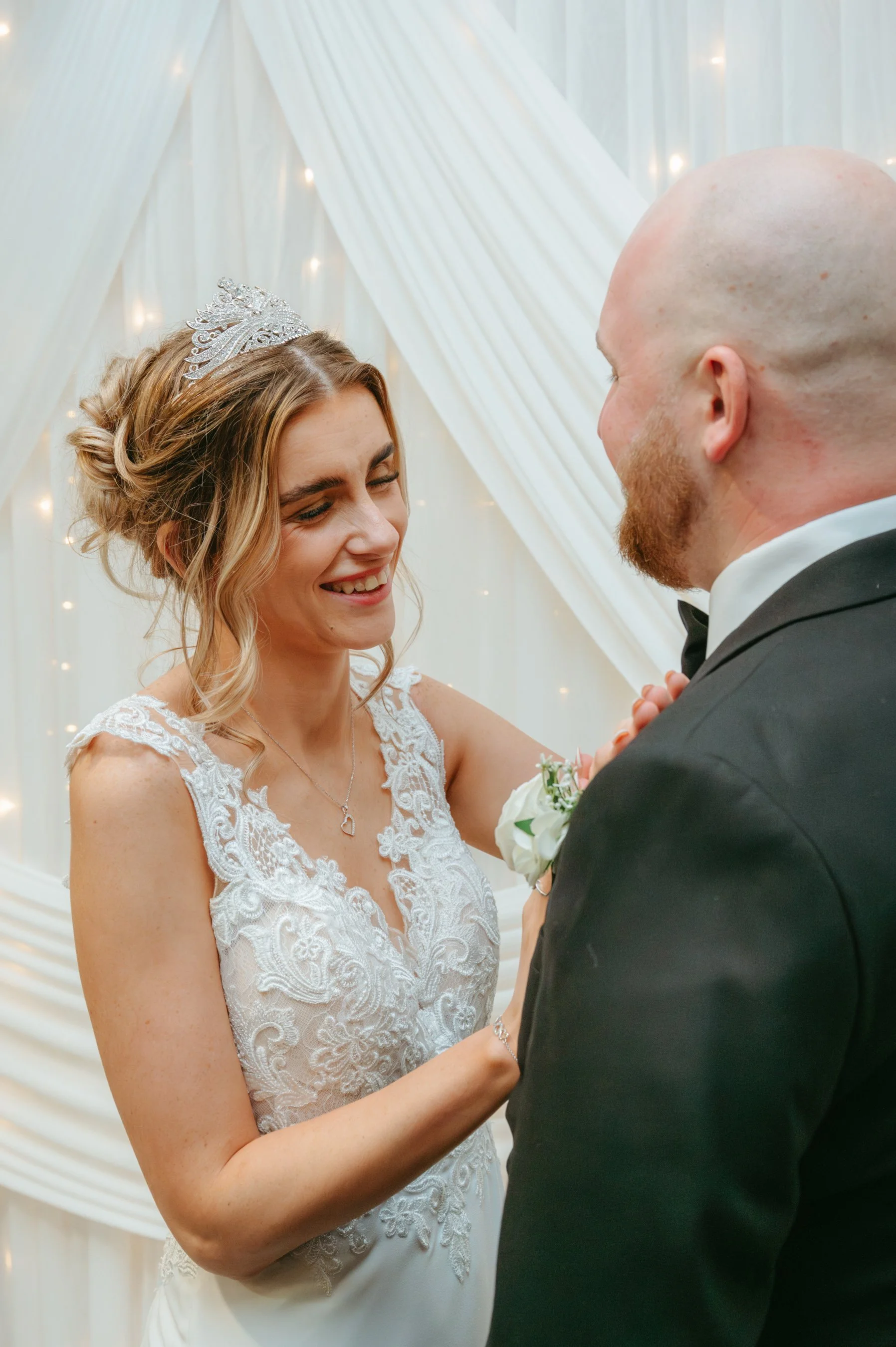 A bride and groom during their wedding ceremony, smiling at each other, with a white decorated backdrop with fairy lights.