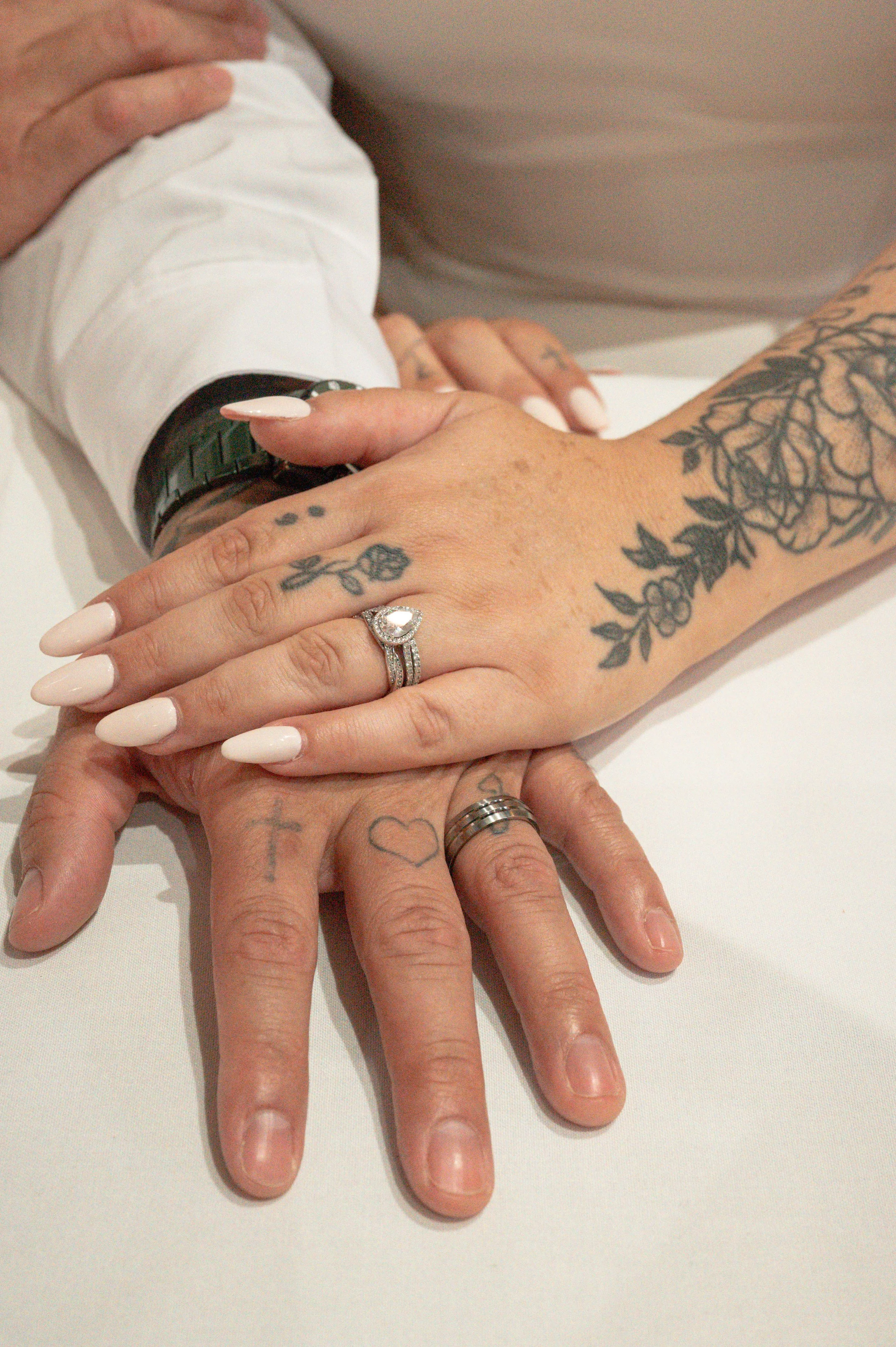 Two hands with tattoos and rings, one with a wedding ring, resting on a white table, with a white long-sleeve shirt visible on the arm. The hands are intertwined or overlapping.