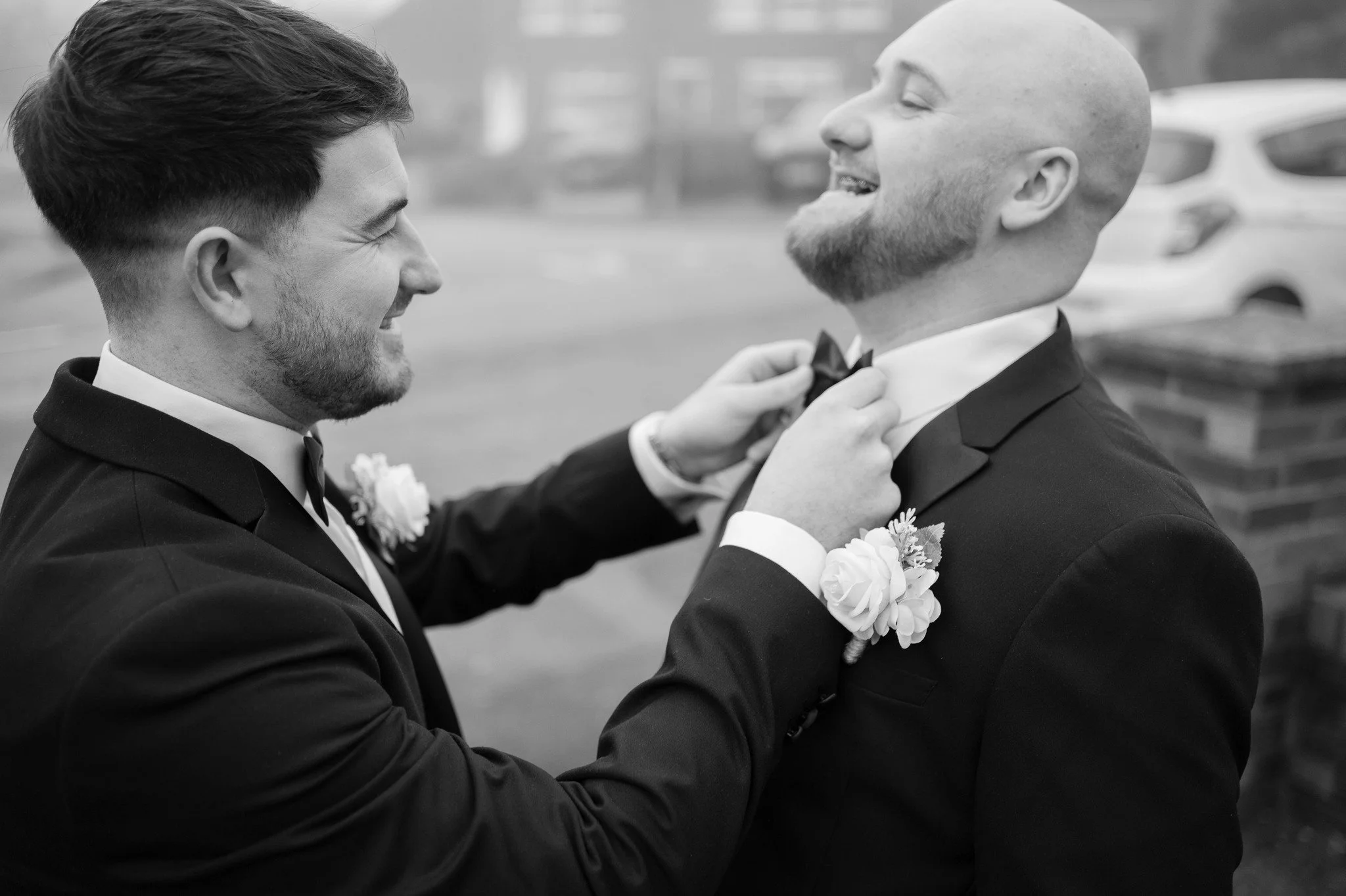 Two men in tuxedos, one adjusts the other's bow tie, both smiling, outdoor setting in black and white.