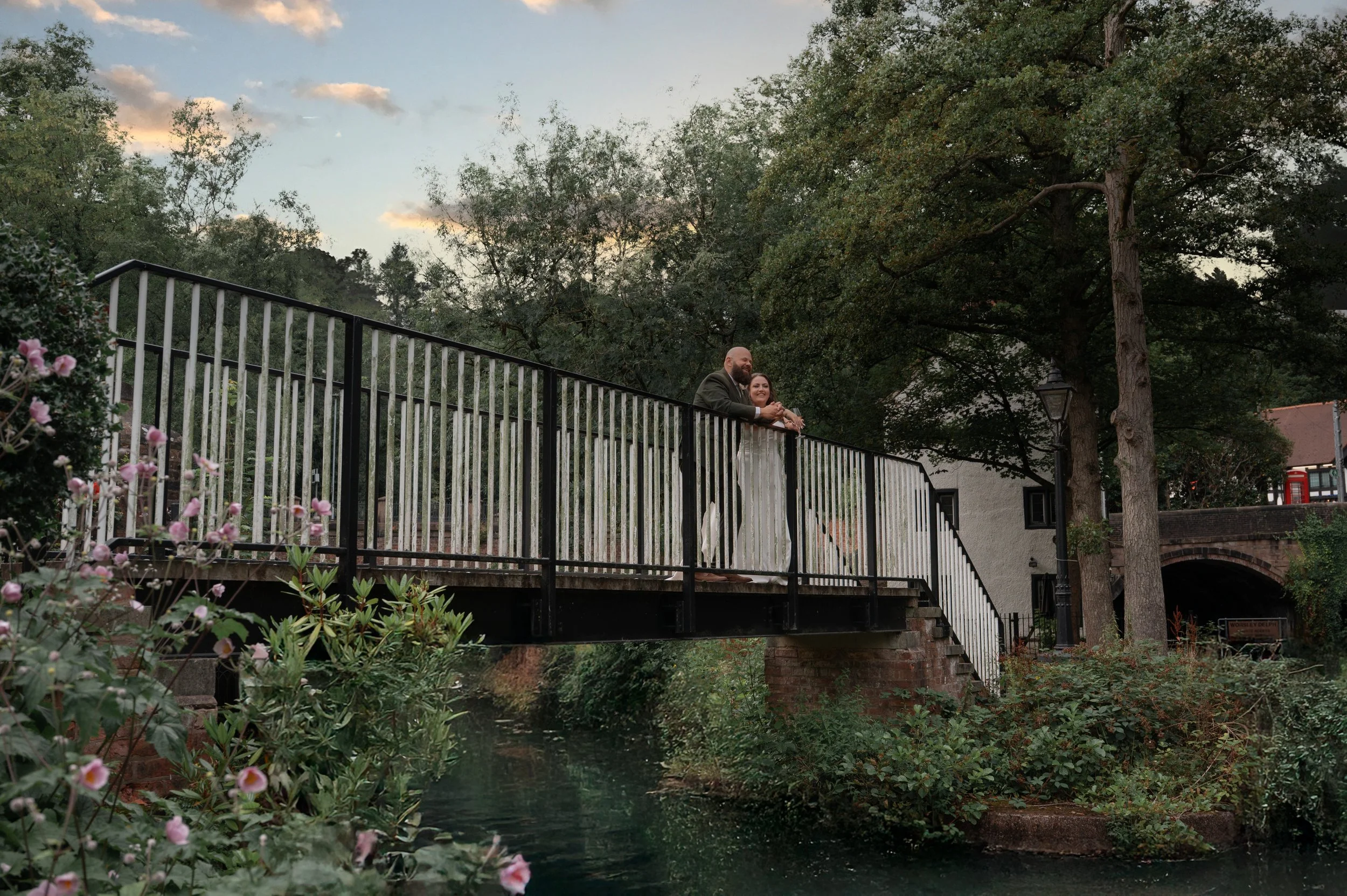 A couple dressed in wedding attire leaning on a black railing bridge over a small creek with trees and buildings in the background.