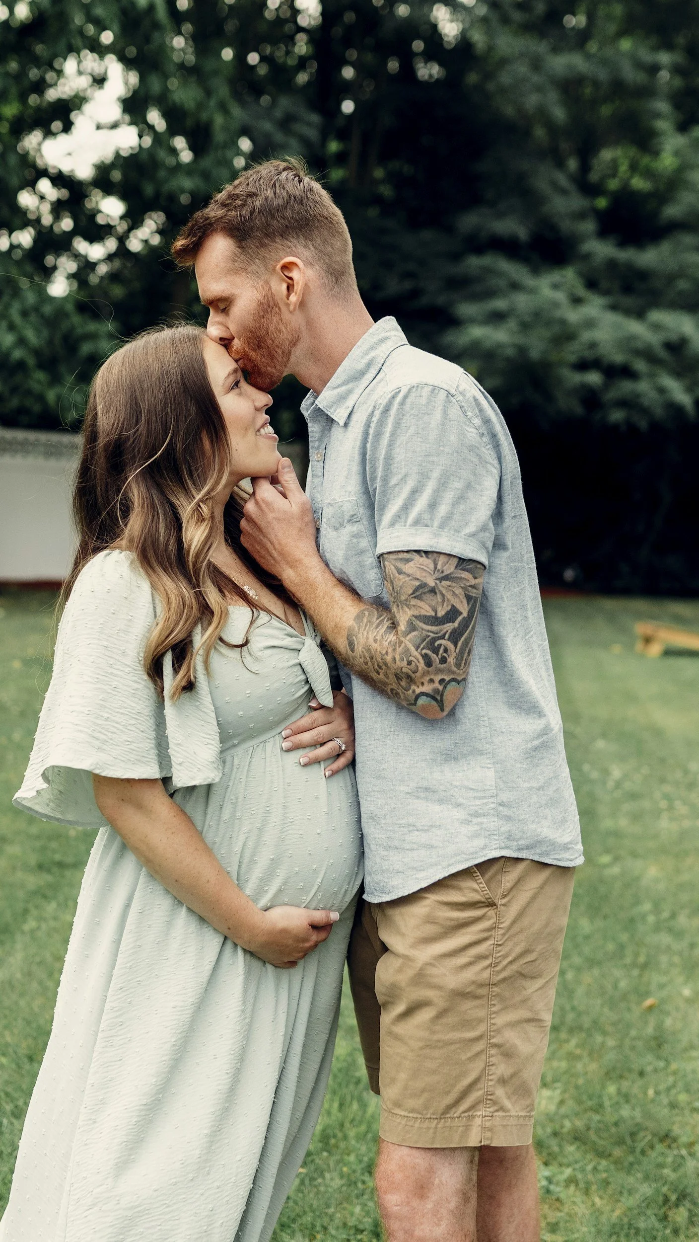 A pregnant woman and a man standing outdoors in a park, smiling and touching her belly, with the man kissing her forehead.