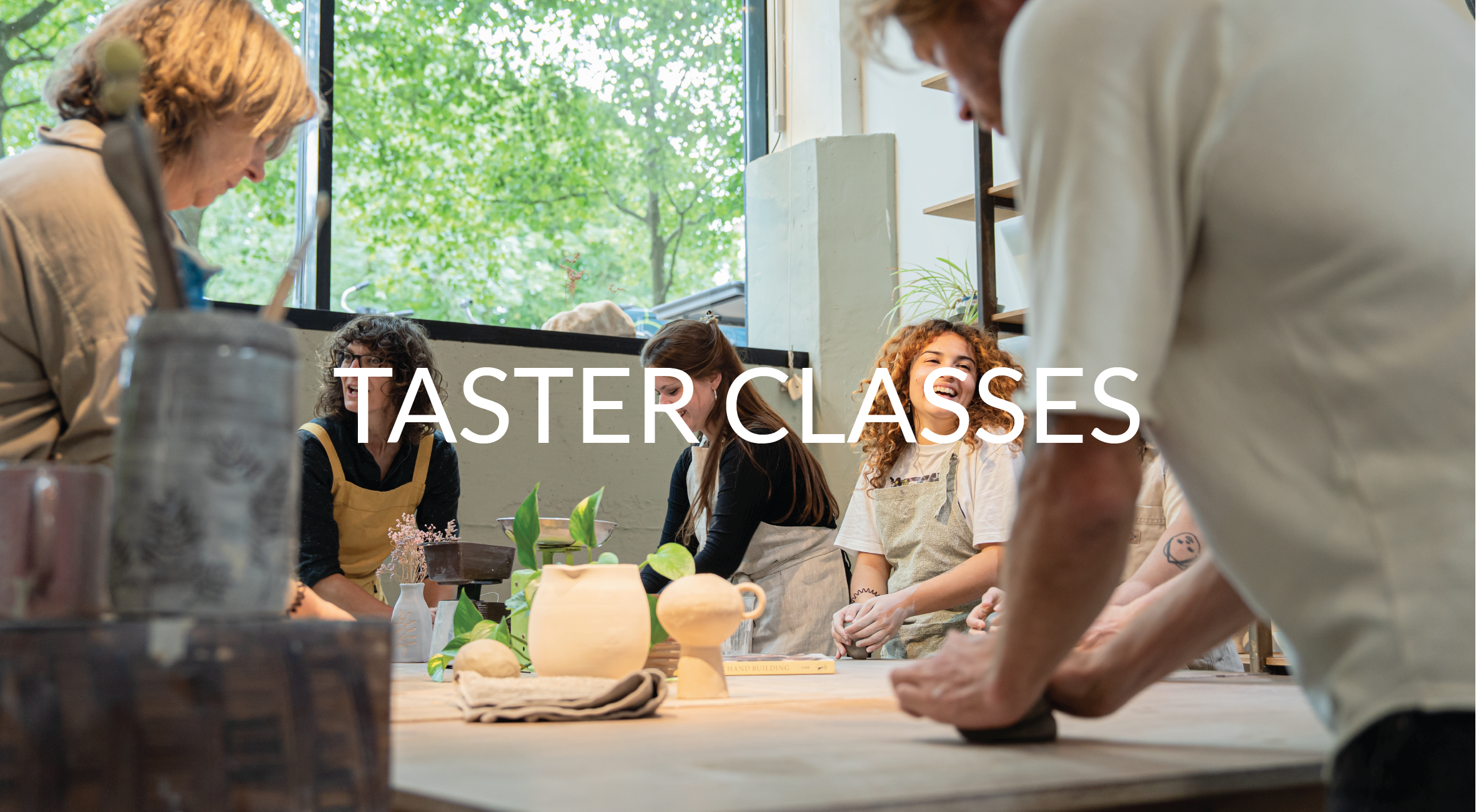 Group of people participating in a pottery or ceramic class, working on clay projects at a workshop table in a studio with large window and green trees outside.