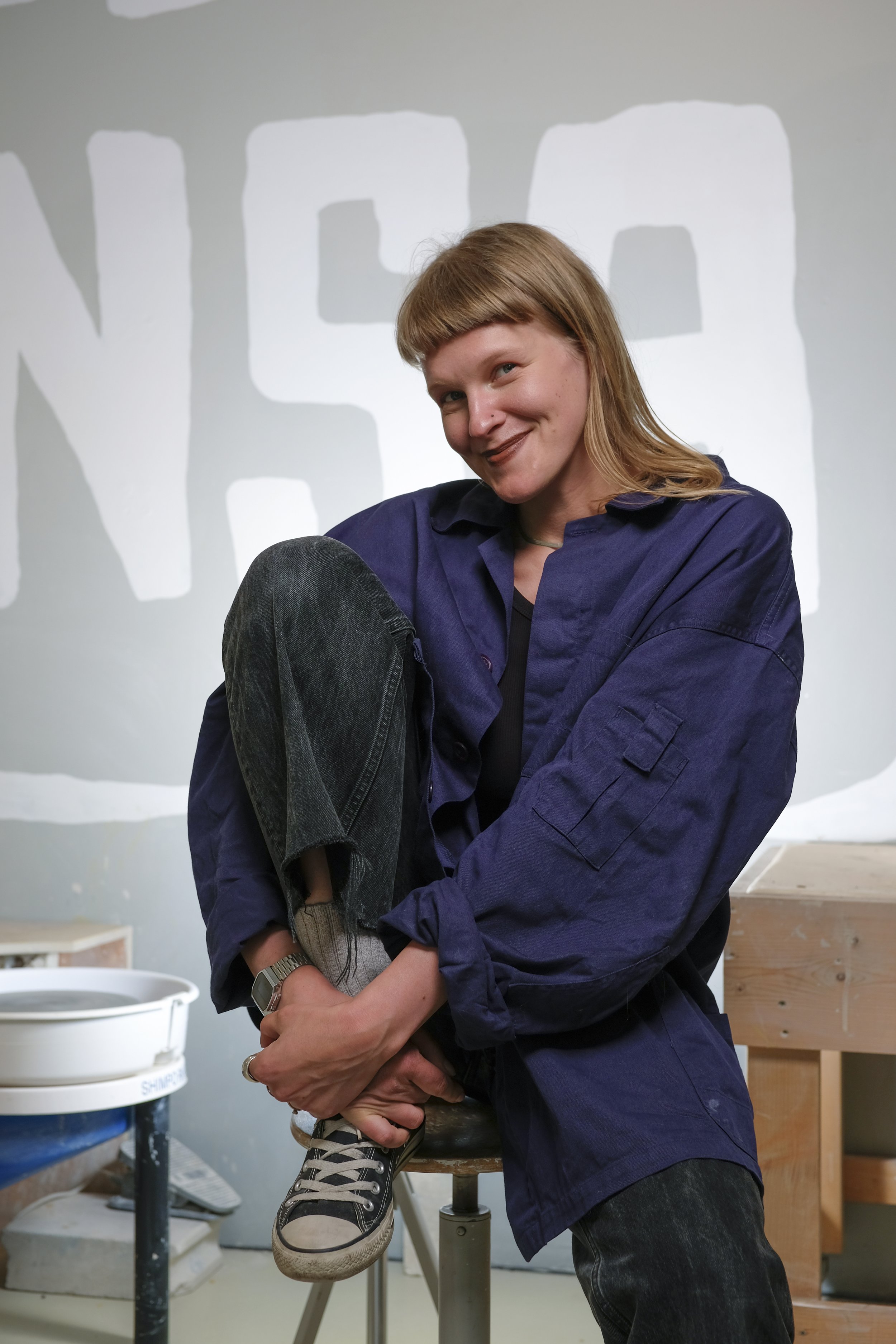 A woman with reddish-brown hair, wearing a navy blue jacket, sitting on a stool with one leg folded on her knee, smiling in front of a painted wall. She is wearing black jeans, gray sneakers, and a watch on her wrist.