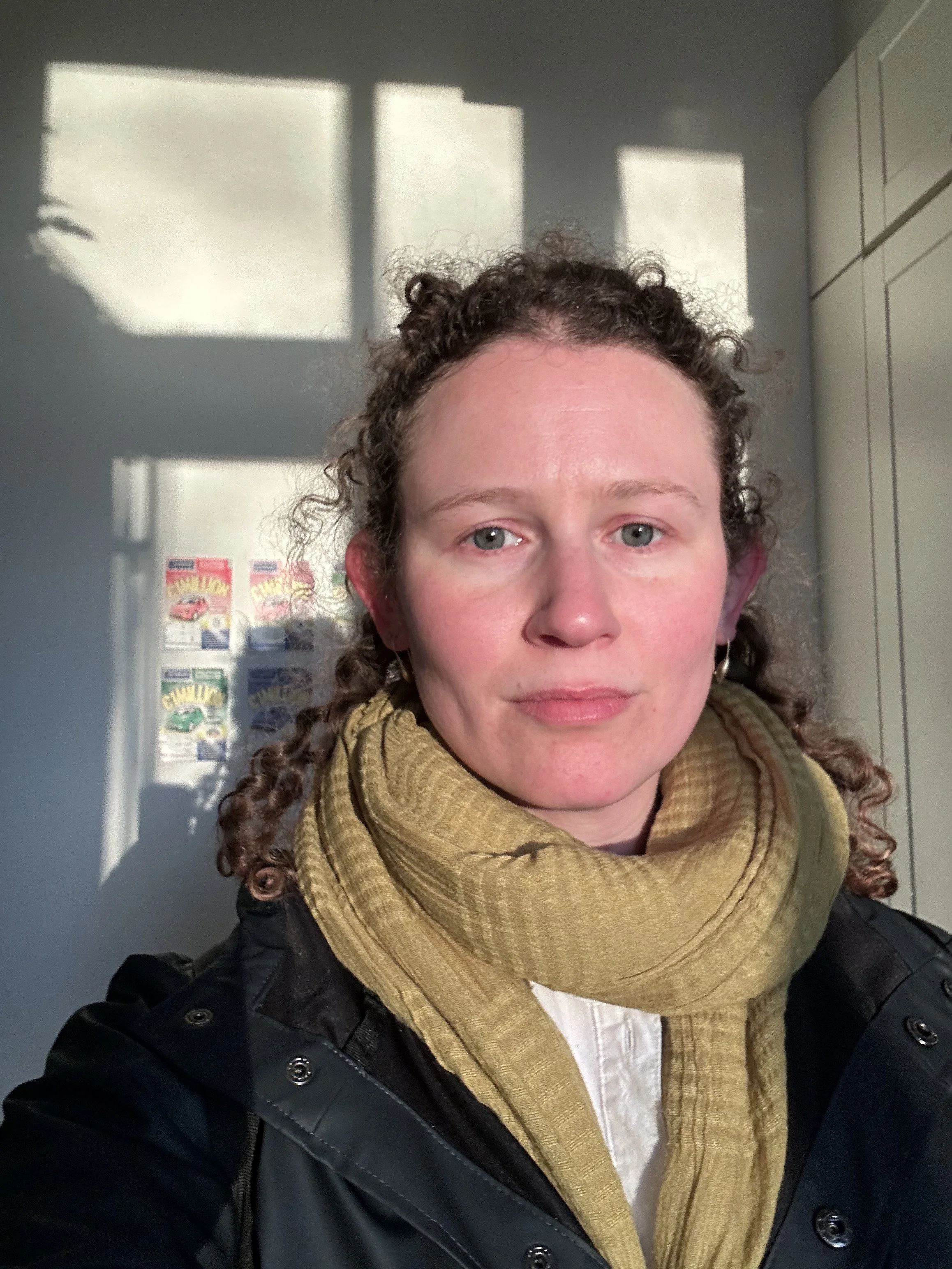 A woman with curly hair wearing a black jacket and a yellow scarf takes a selfie in a room with sunlight casting shadows on the wall behind her.