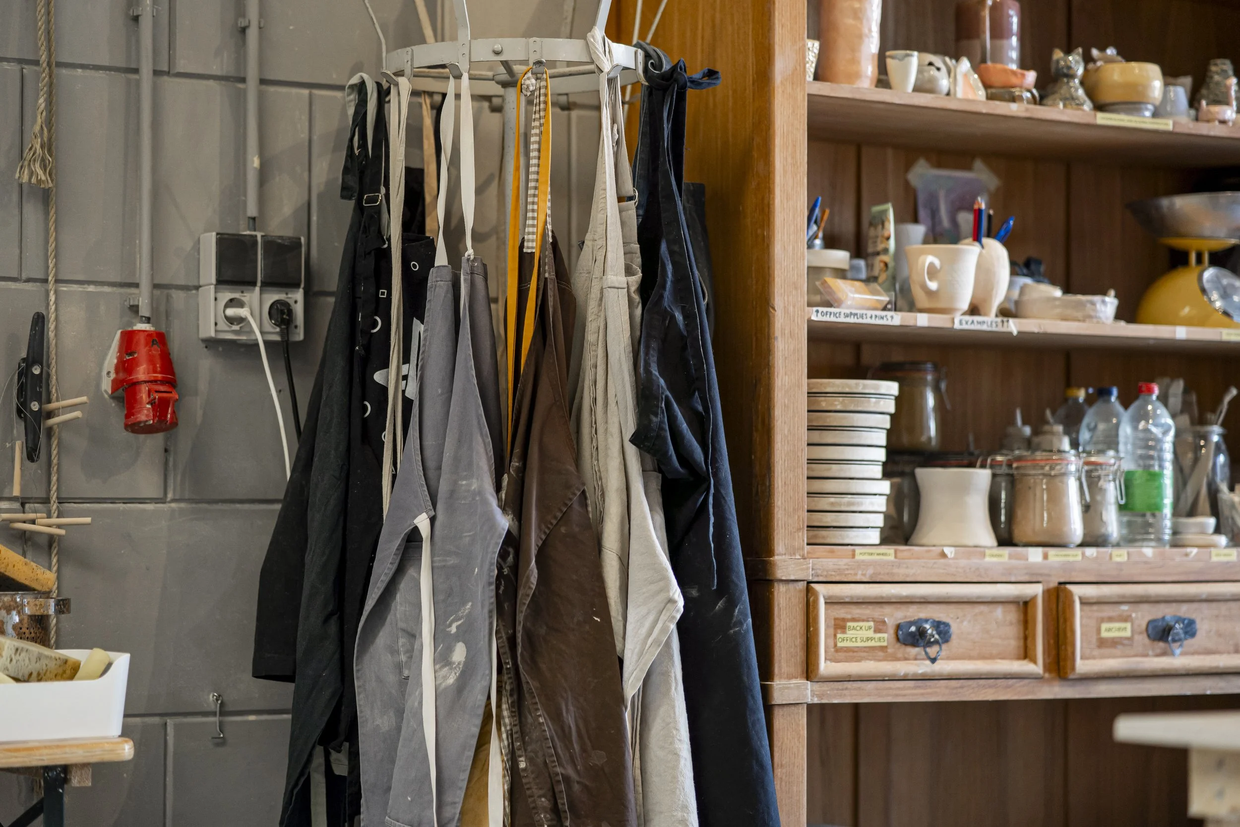 A clothing rack with aprons hanging next to a wooden shelf filled with dishes and kitchen supplies.