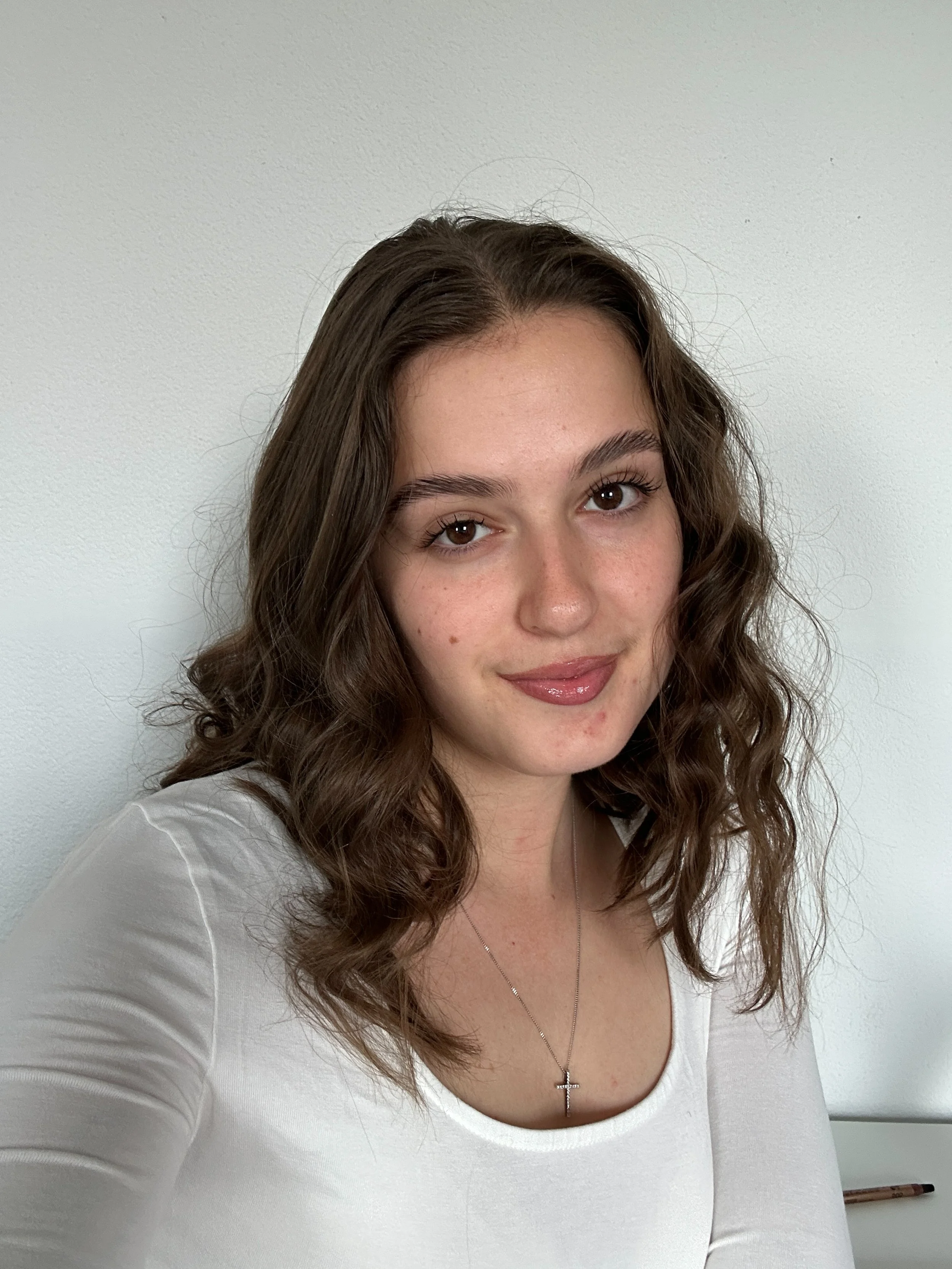 A young woman with wavy brown hair, wearing a white top and a cross necklace, smiling softly and looking at the camera, against a plain white wall background.