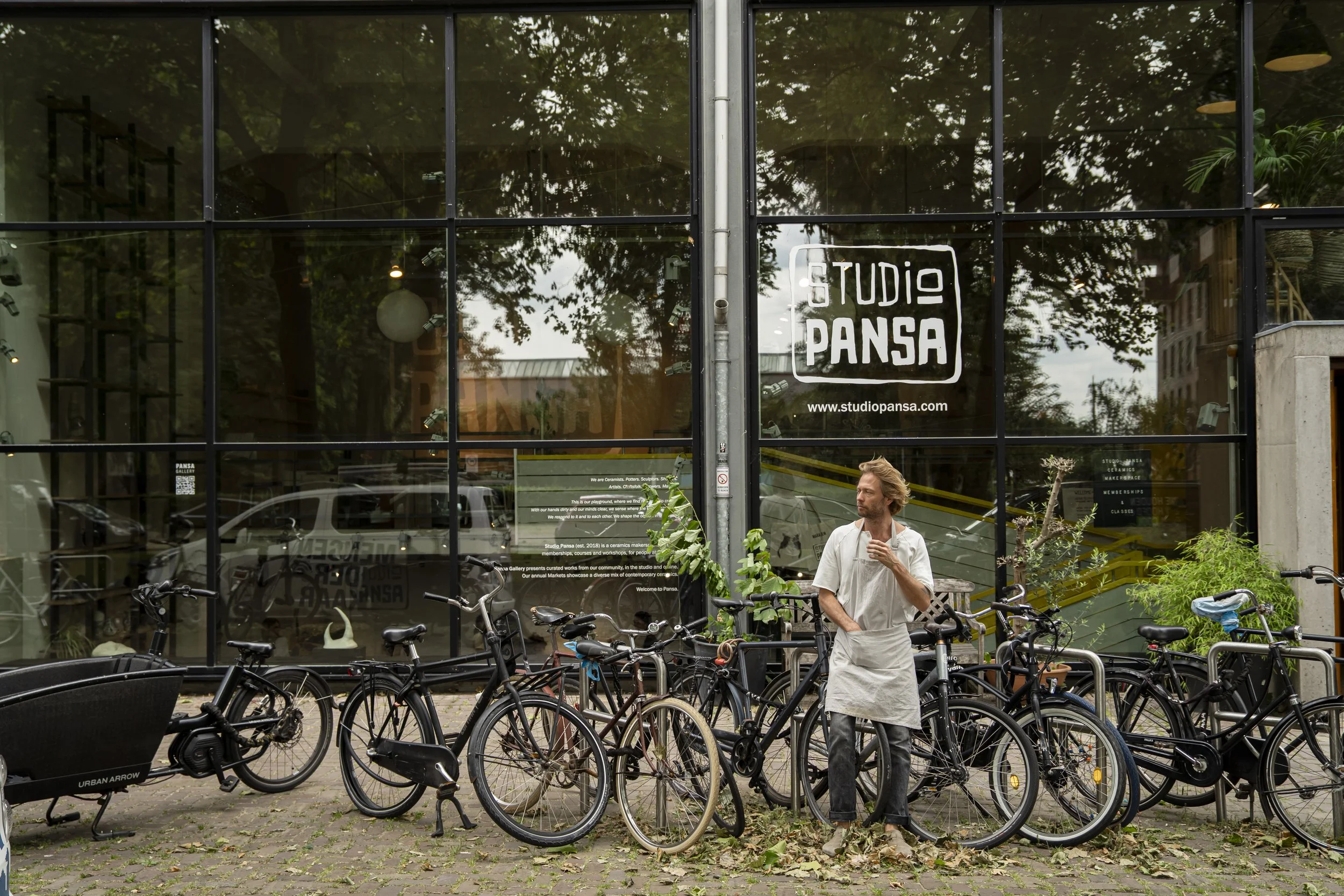 A man stands beside bicycles outside Studio Pansa, a building with large glass windows. The man is wearing a white shirt and apron, holding a drink, and looking to the side. The storefront has the studio's logo and website on the window.