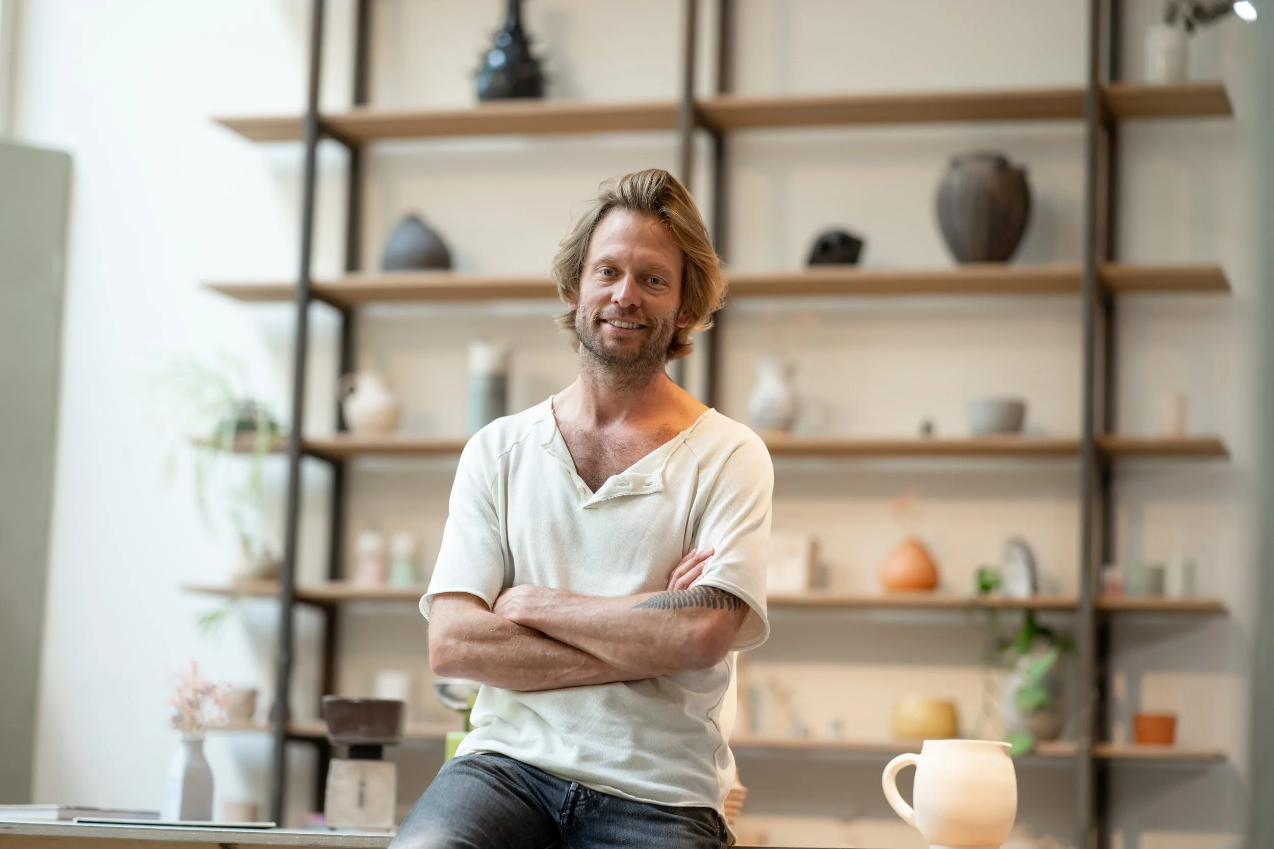 A man with light brown hair and a beard, smiling with arms crossed, standing in a pottery studio with wooden shelves filled with ceramic vases, bowls, and plants in the background.