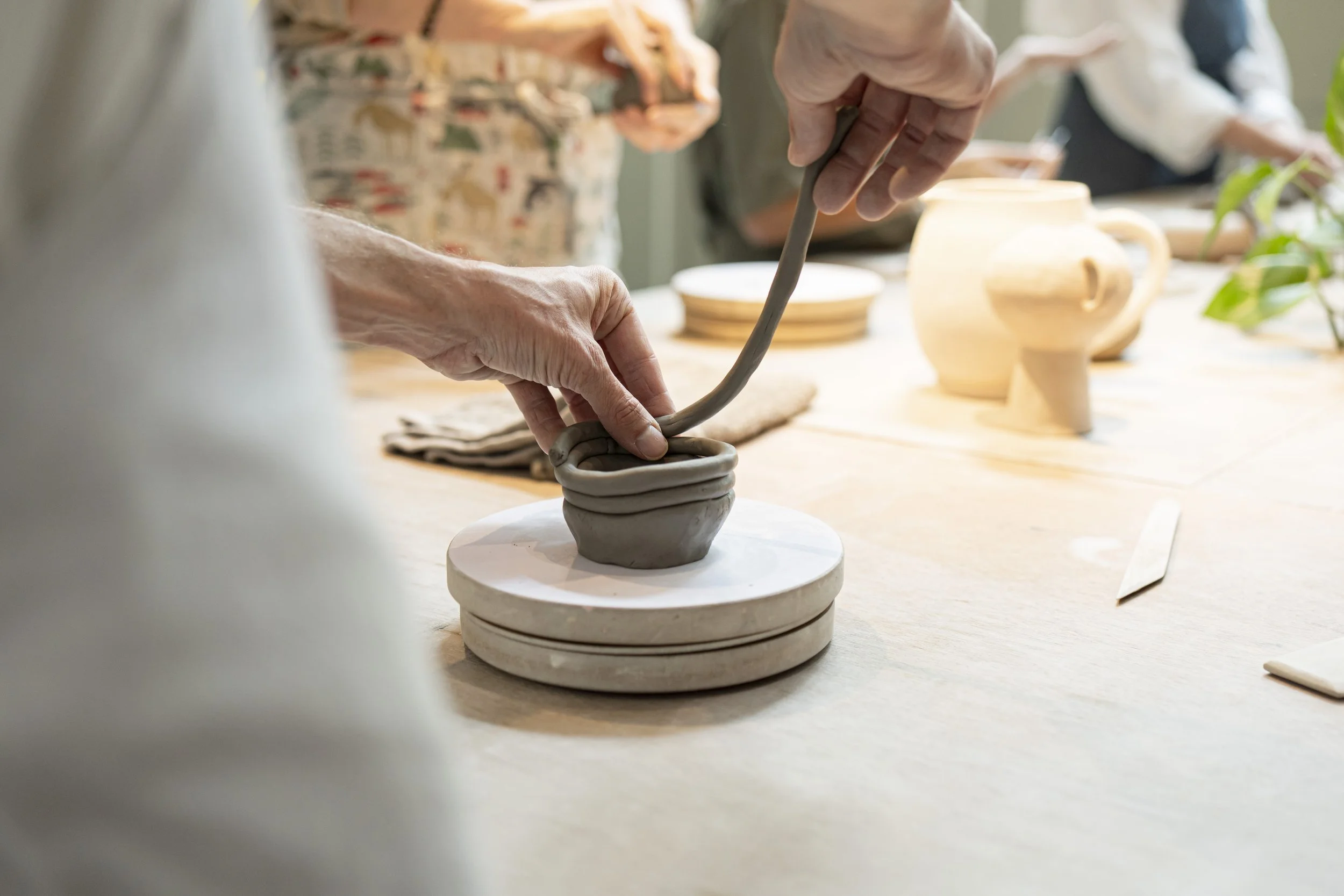 People shaping and decorating pottery on a table in a ceramics class or workshop, with unfinished ceramic pieces and plates in the background.