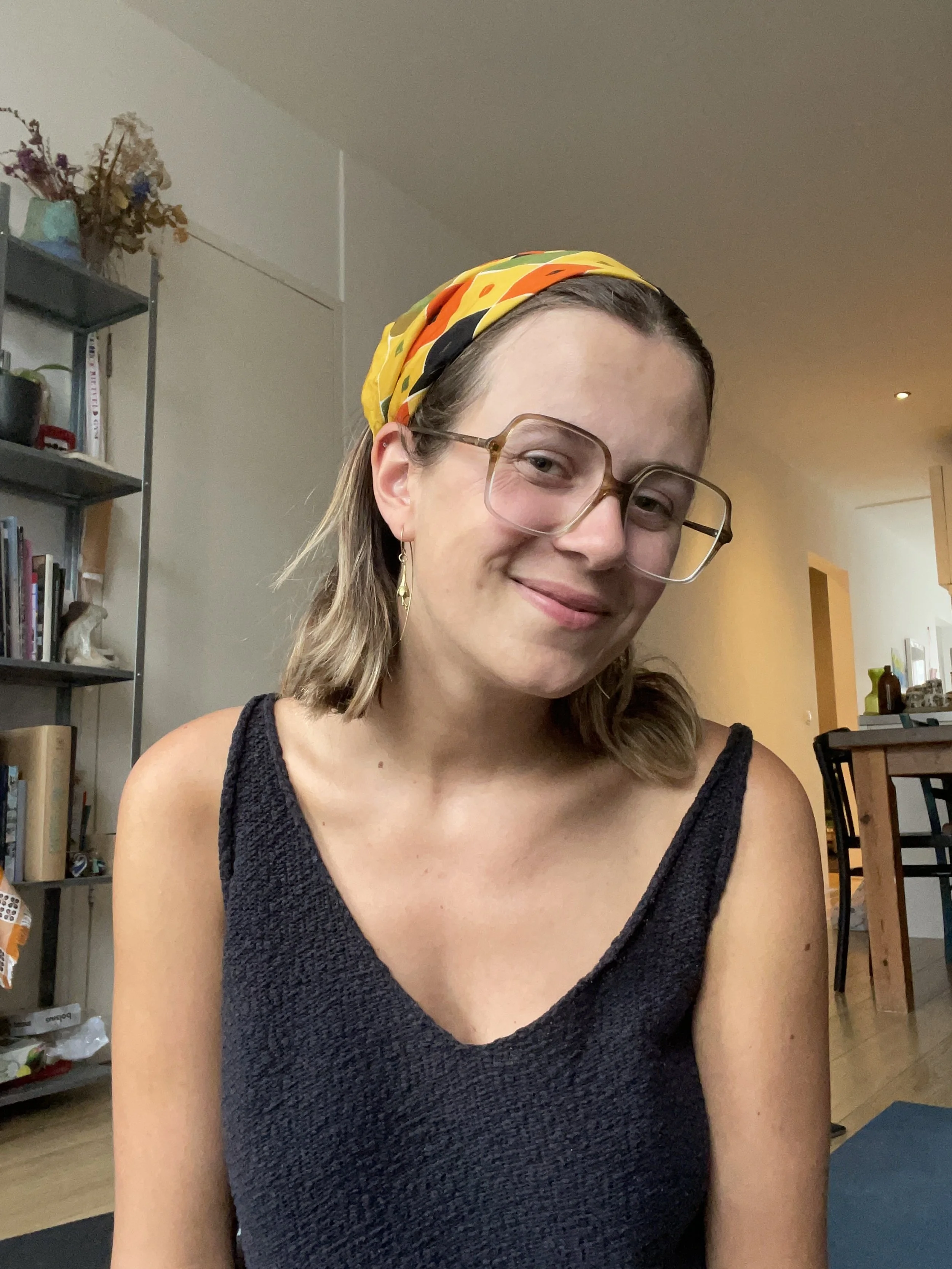A woman with short blonde hair, wearing glasses, a yellow headscarf with orange and black patterns, earrings, and a sleeveless dark top, smiling softly in a cozy indoor setting with bookshelves and dining furniture in the background.