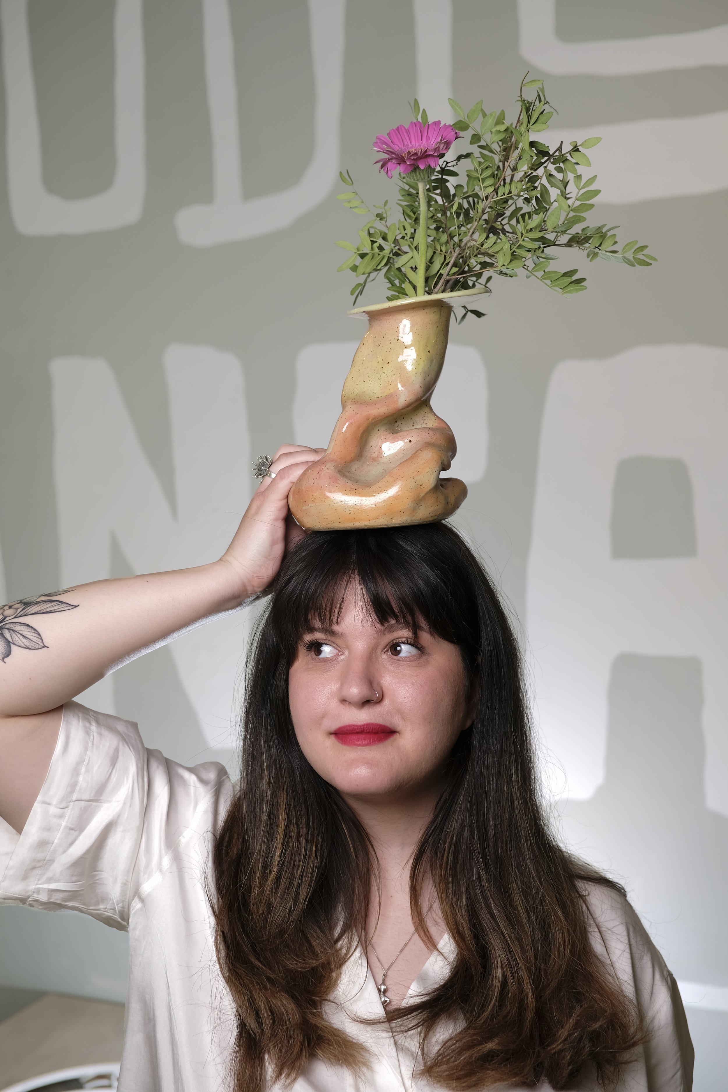 A woman with dark hair and red lipstick holds a ceramic shoe-shaped vase with pink flowers and green leaves on her head.