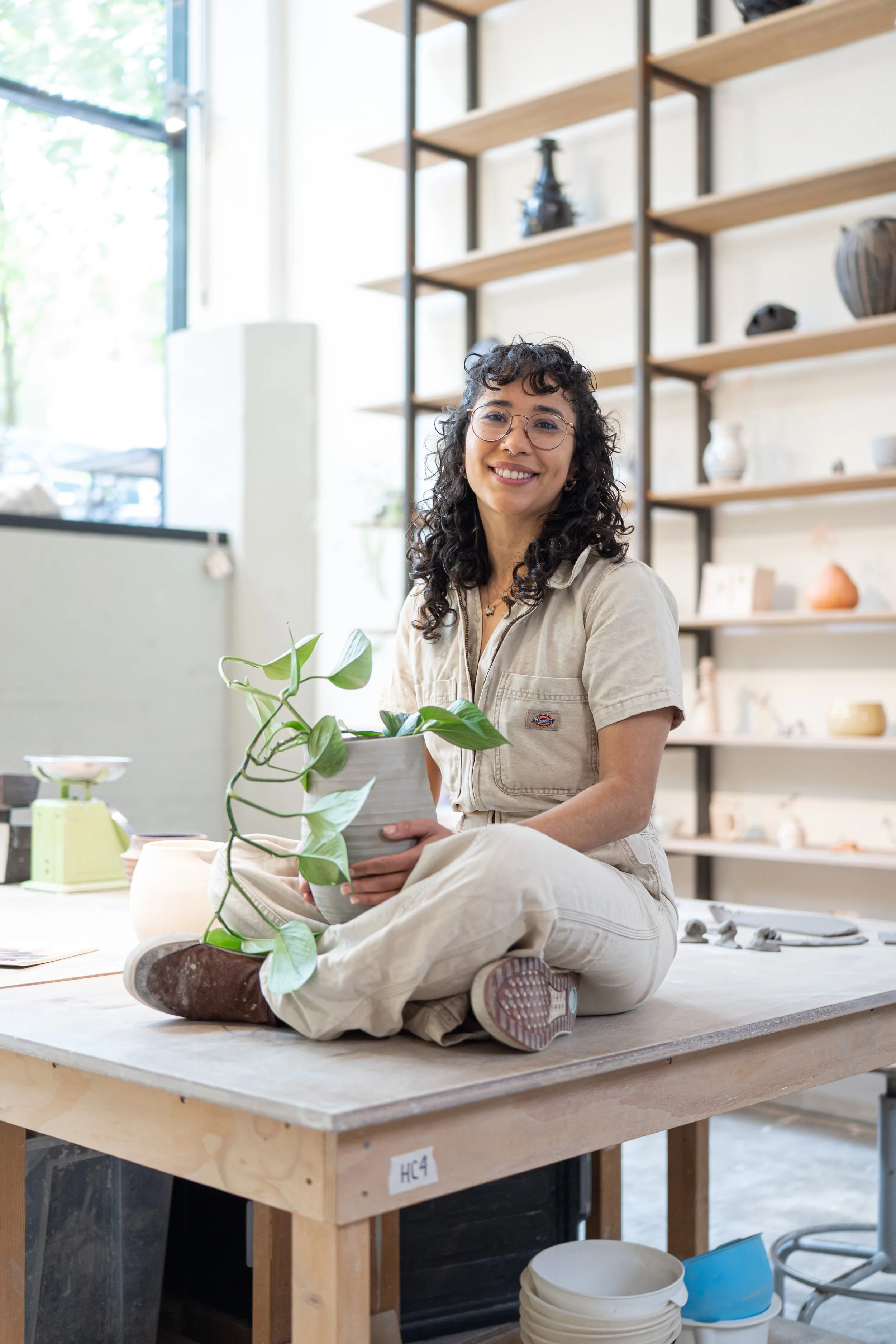 A woman sitting cross-legged on a woodworking table in a studio, holding a potted plant, smiling at the camera with shelves filled with various pottery and decorative objects in the background.