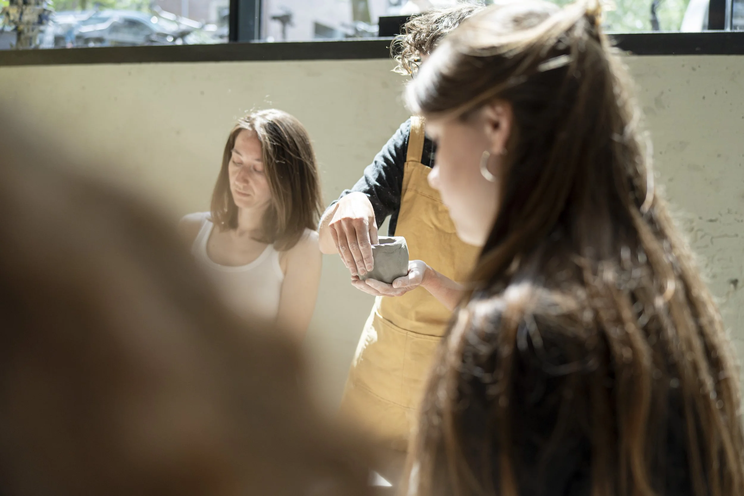 A woman with brown hair and hoops earrings looking at pottery being made by an instructor in a yellow apron, with another woman with brown hair and a tank top in the background.