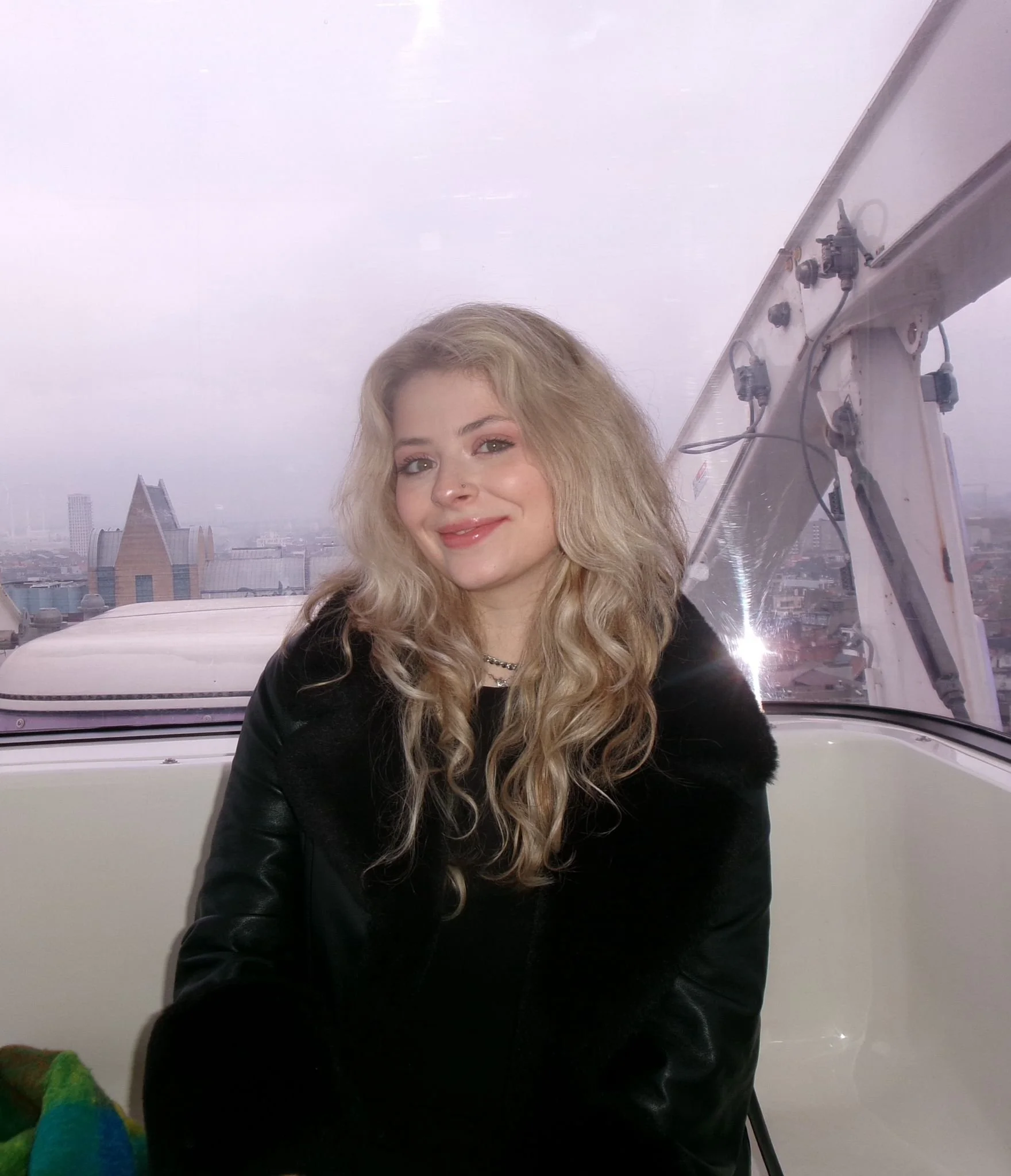 A young woman with long blonde curly hair smiling inside a cabin with a city skyline visible through the window behind her.