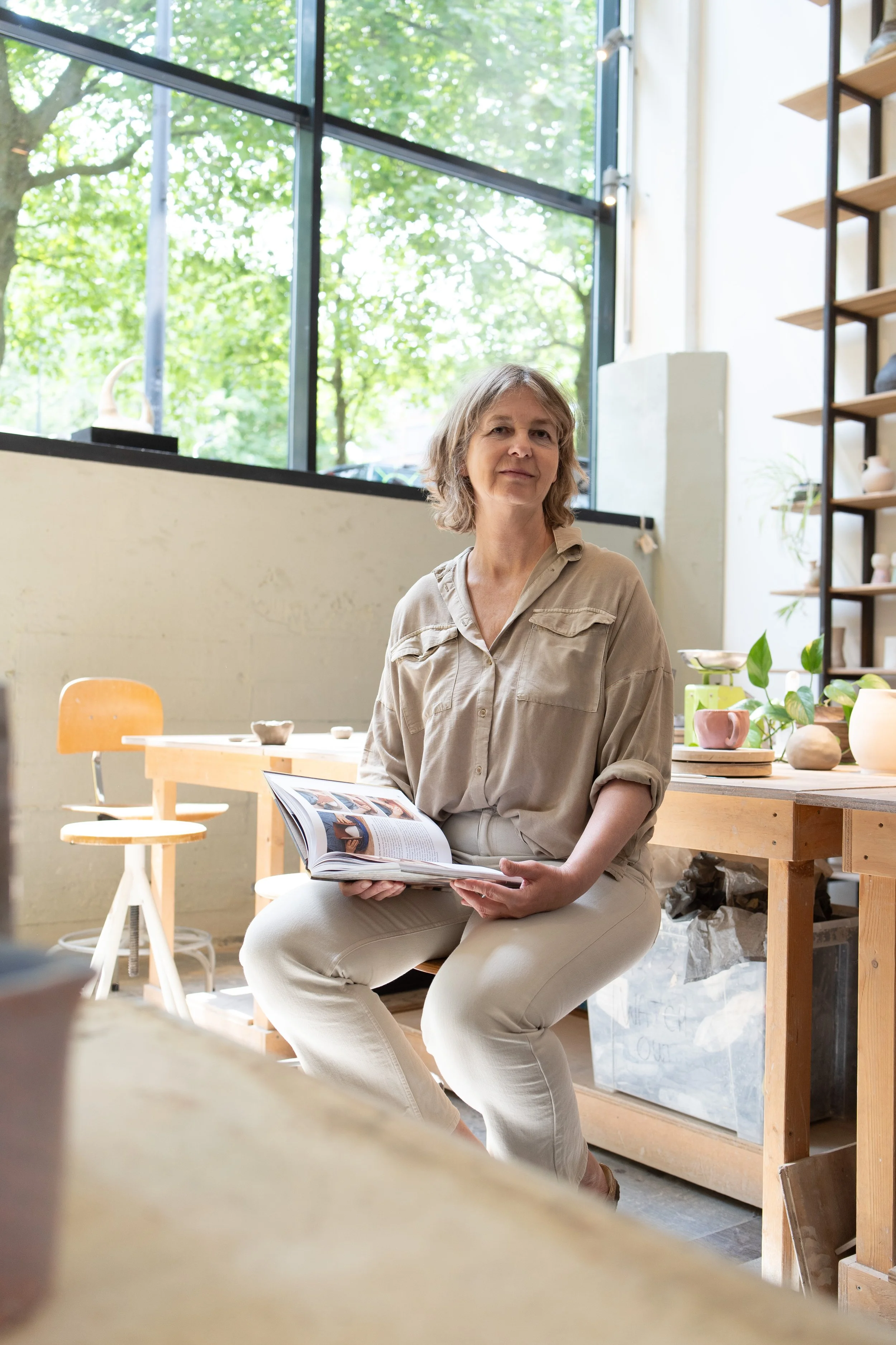 A woman with short wavy blonde hair sitting indoors on a wooden stool, holding an open magazine, in a well-lit room with large windows, wooden shelves with ceramics and potted plants.