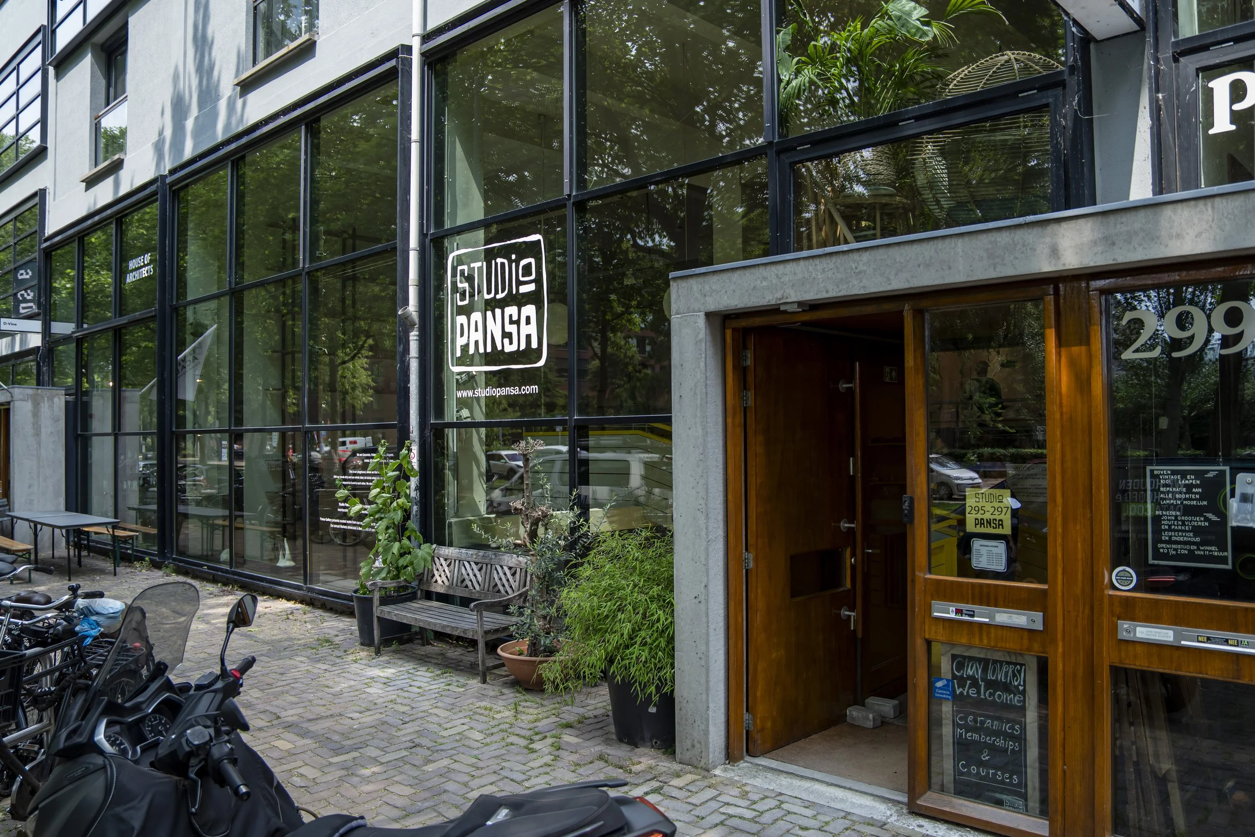 Exterior of an architecture studio named Studio Pansa, with large glass windows, potted plants, benches, and bicycles parked outside. The entrance has a wooden door with signs and a chalkboard.